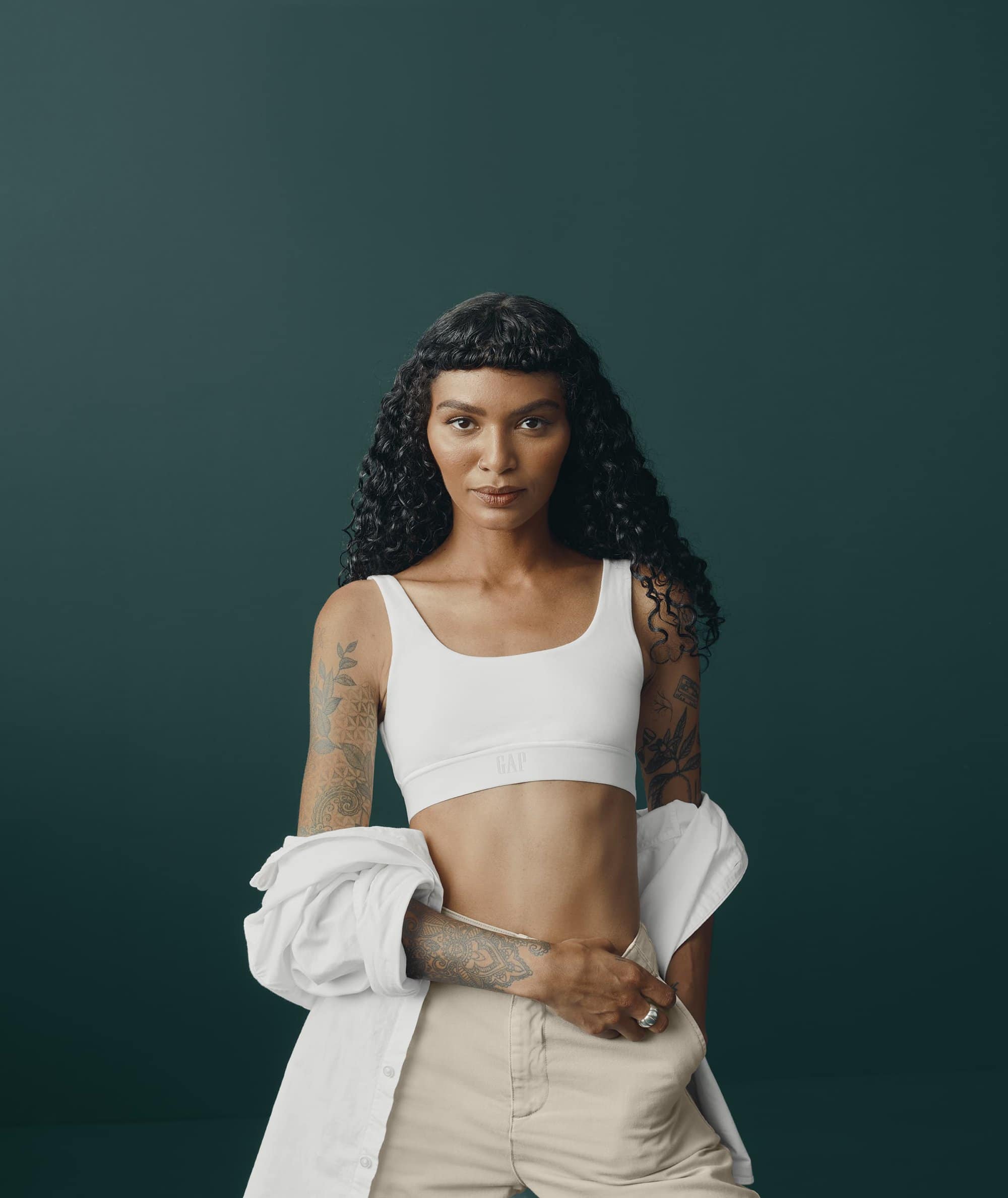 Portrait of a woman with curly hair in a white top and beige pants, standing against a dark green background in a studio setting.
