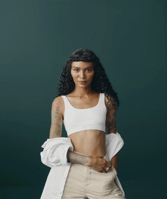 Portrait of a woman with curly hair in a white top and beige pants, standing against a dark green background in a studio setting.