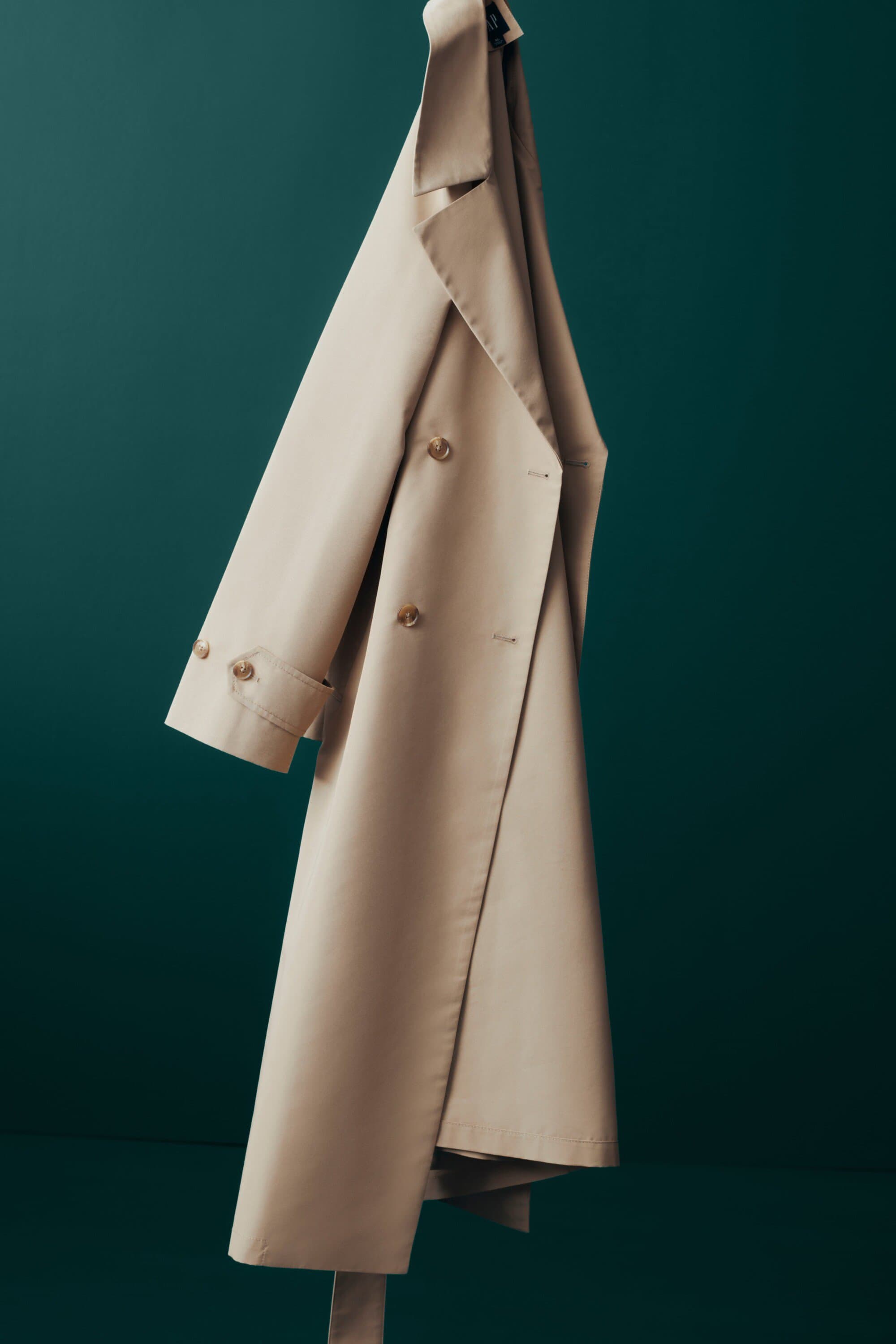 Beige trench coat on hanger against green backdrop in a studio setting, showcasing its double-breasted design and tied waist belt.