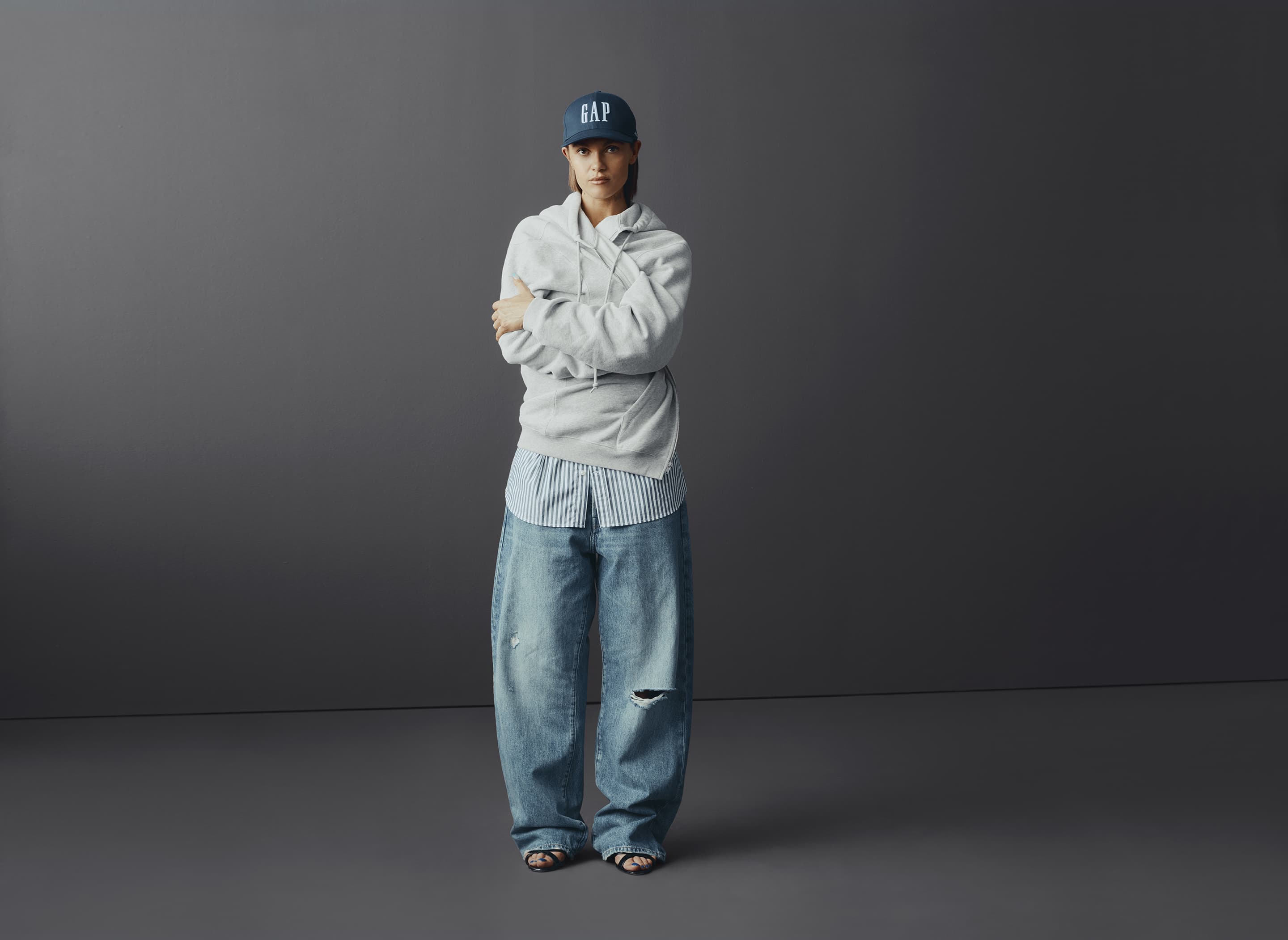 Full-length shot of a young adult woman in a baseball cap, hoodie, blue jeans, and sandals standing against a neutral grey background.