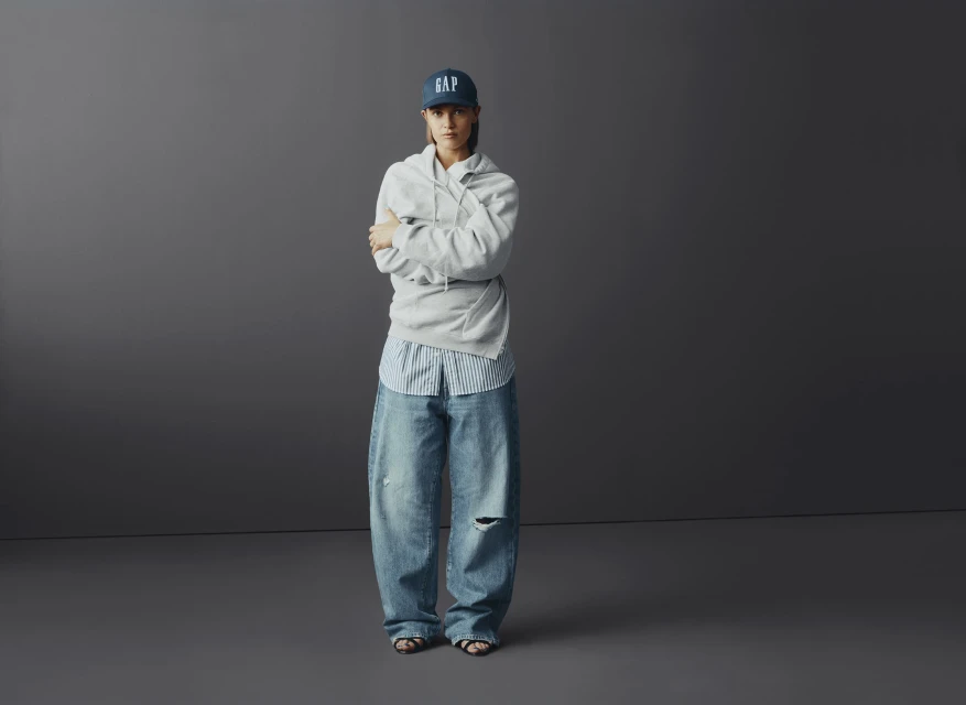 Full-length shot of a young adult woman in a baseball cap, hoodie, blue jeans, and sandals standing against a neutral grey background.