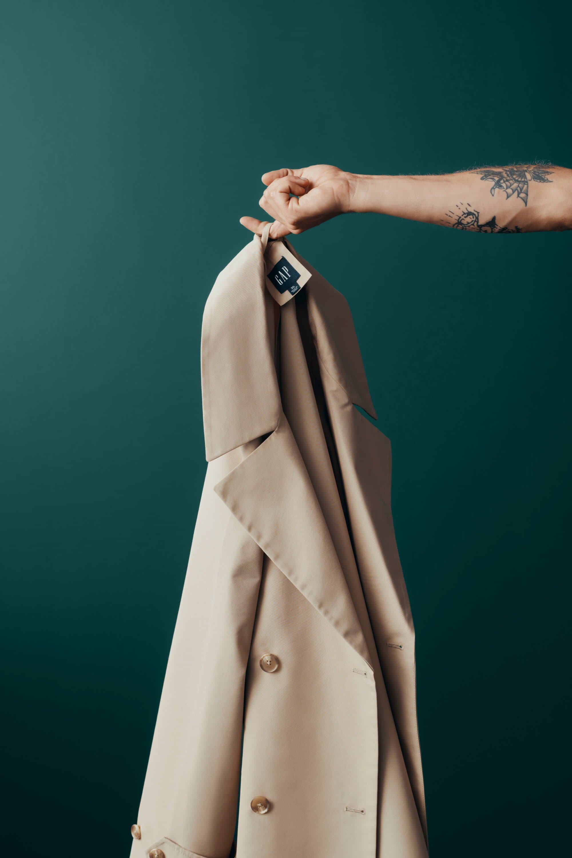 Tan trench coat held up by an arm against a dark teal background, featuring a clear view of the garment's texture and design.