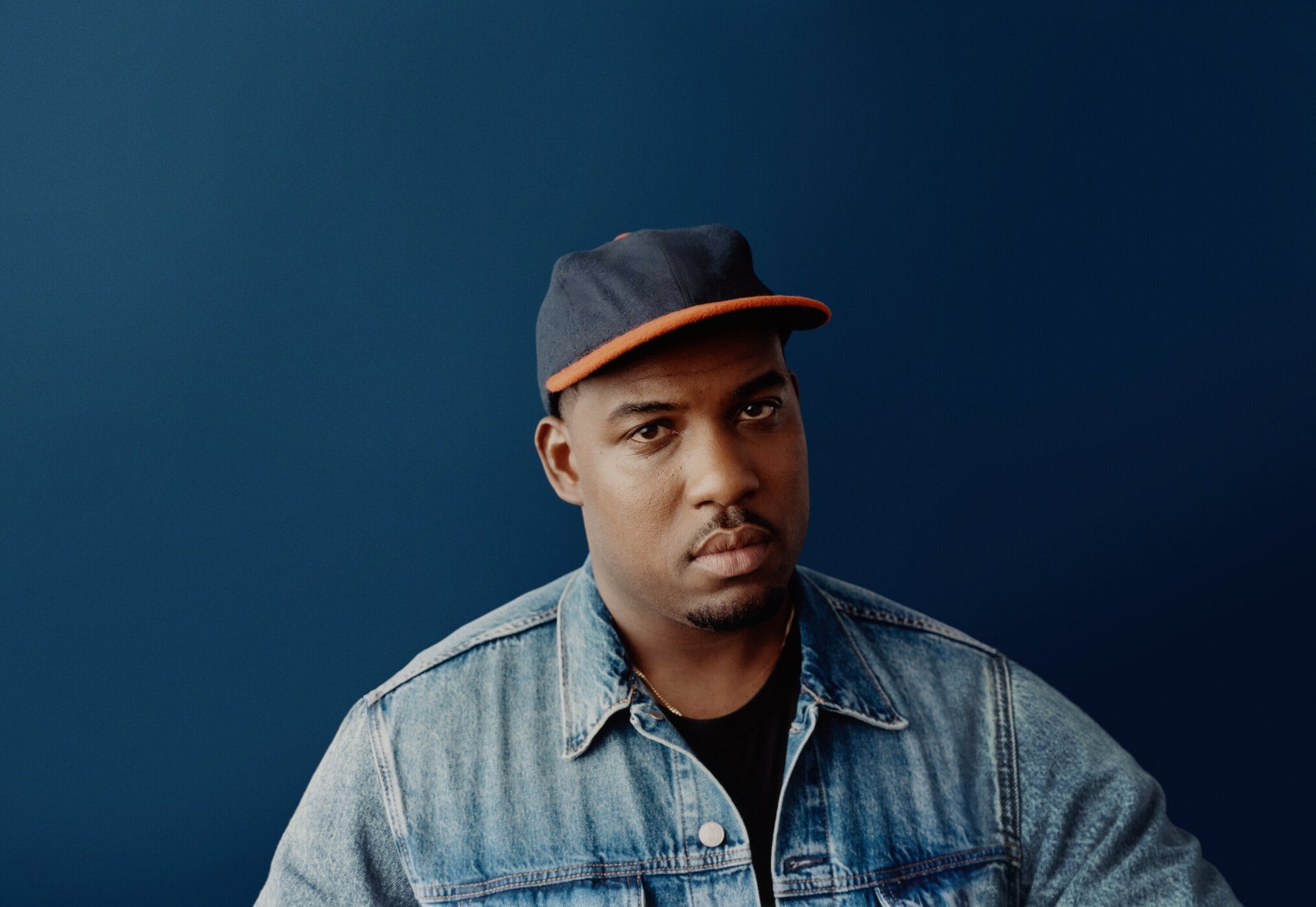 Portrait of a young adult Black man wearing a denim jacket and baseball cap against a dark blue backdrop in a photo studio.