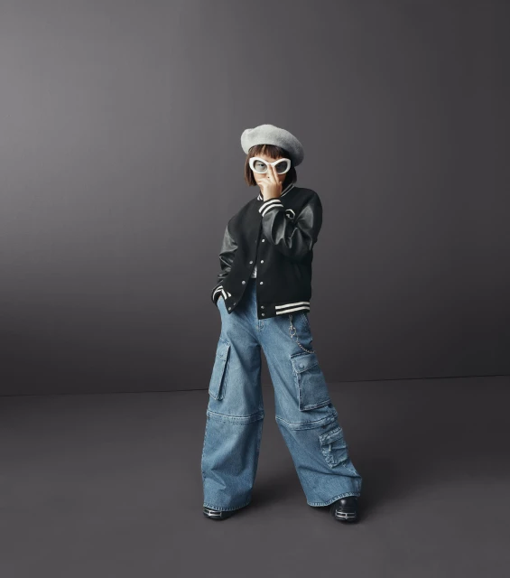 Child model in a black varsity jacket, oversized denim cargo pants, beret, and sunglasses poses in a studio with a neutral background.