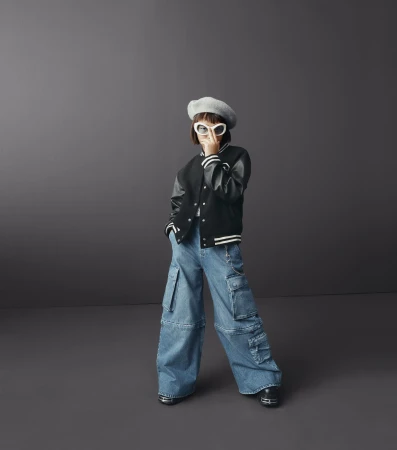 Child model in a black varsity jacket, oversized denim cargo pants, beret, and sunglasses poses in a studio with a neutral background.
