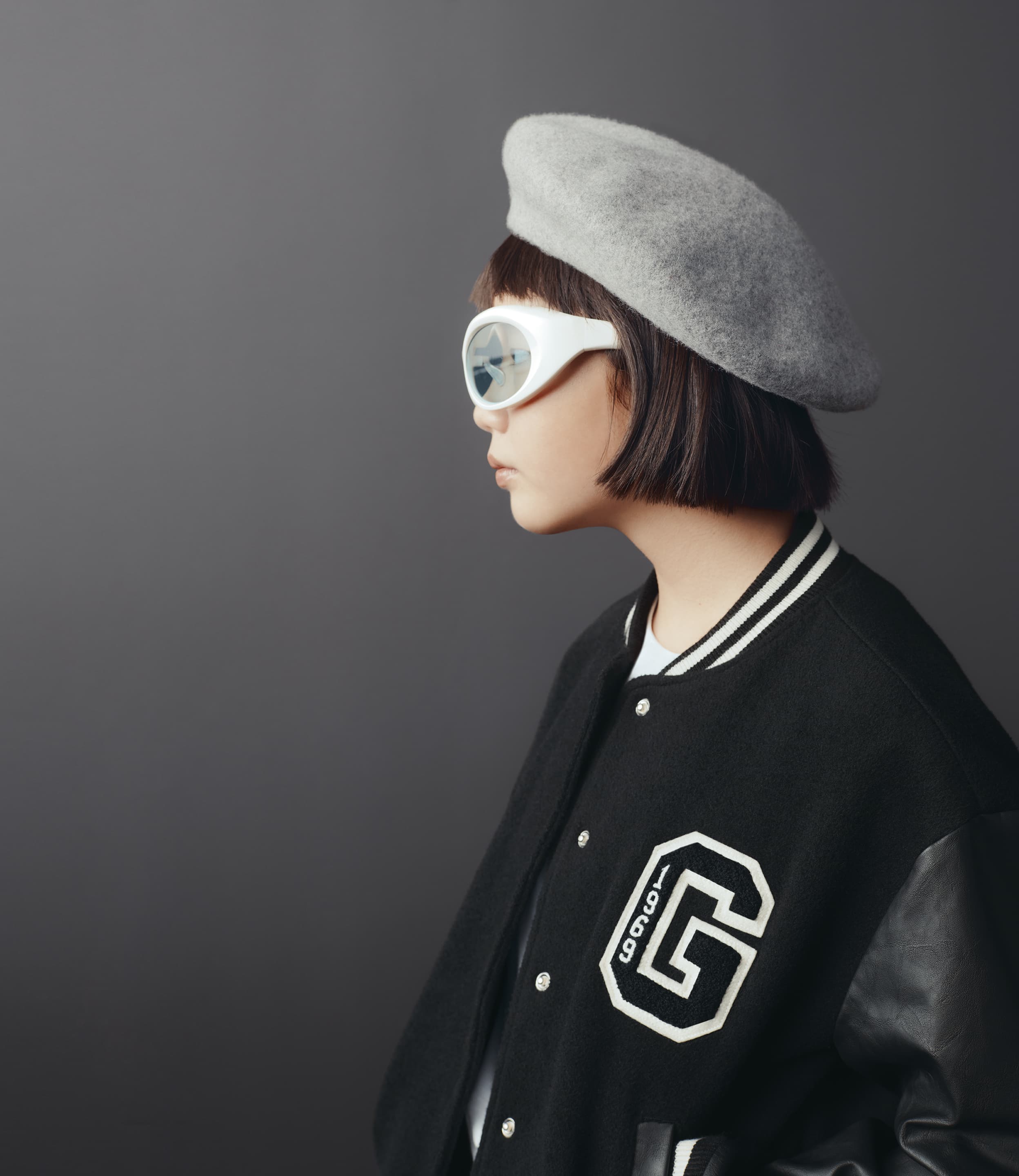 Young woman in a beret and jacket with retro sunglasses poses in front of a neutral gray studio backdrop, side view, fashion portrait.