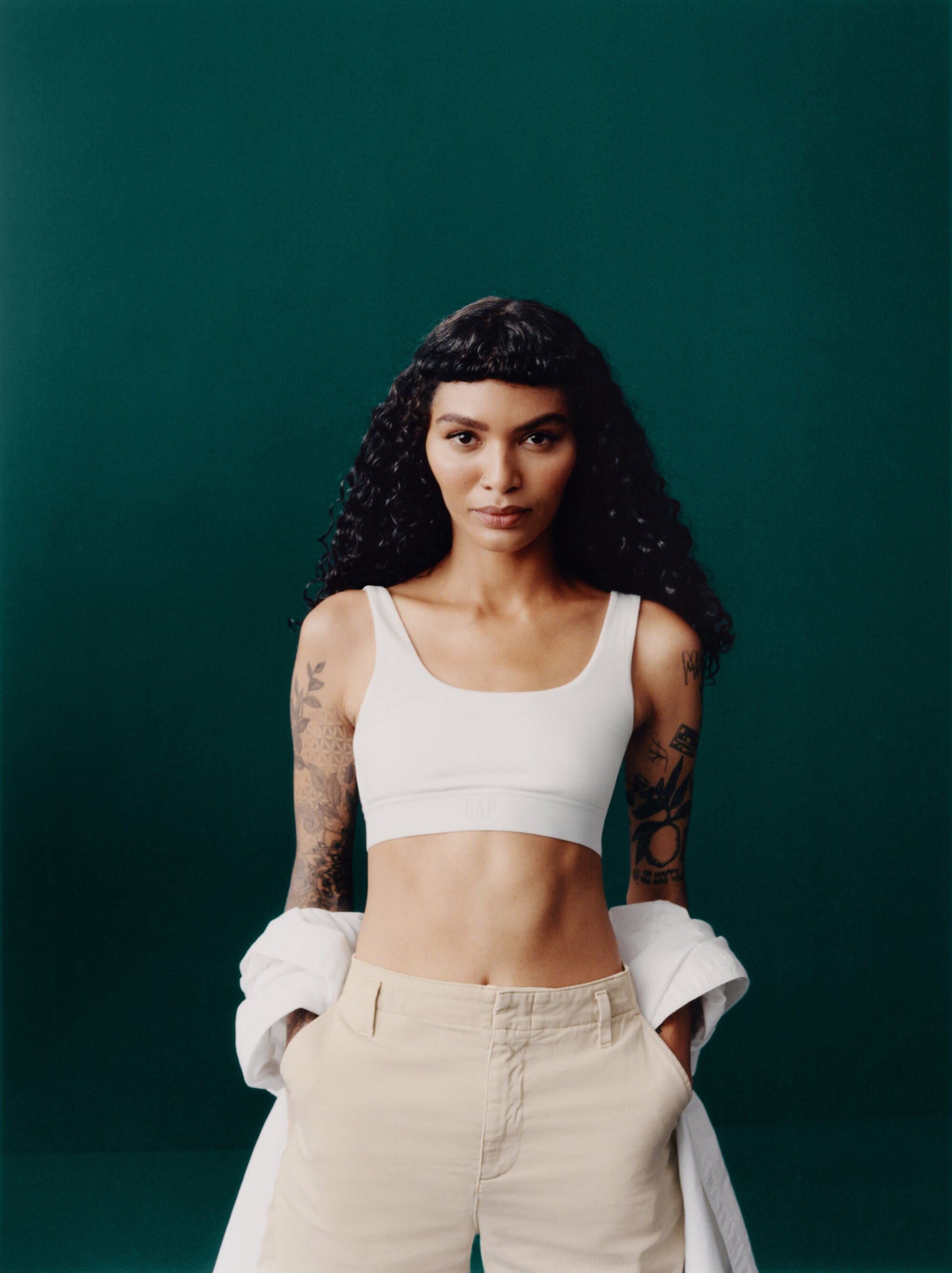 A woman with dark curly hair and tattoos poses in a white top and tan shorts against a dark green background in a studio setting.