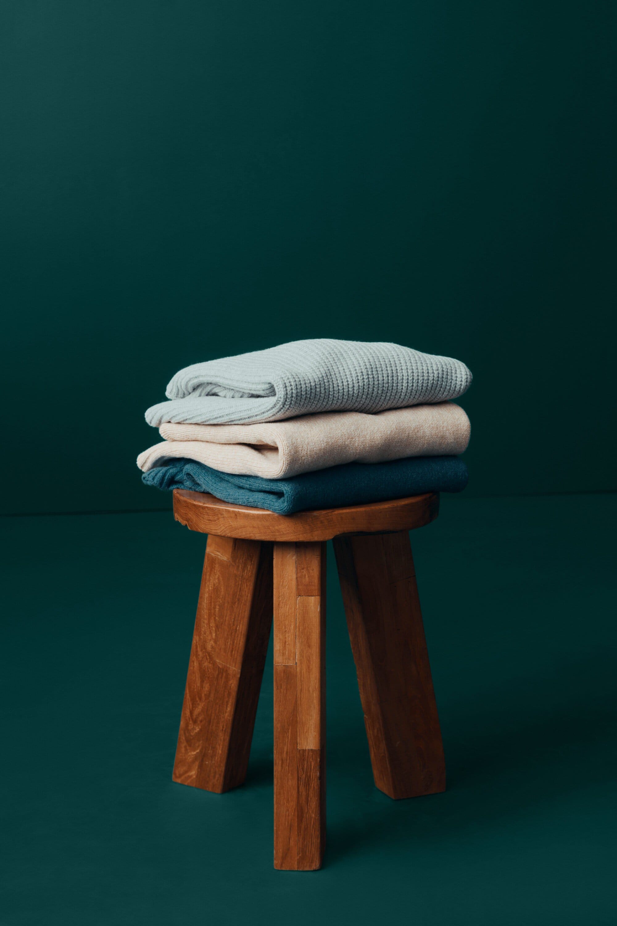 Stack of folded knit sweaters on a wooden stool against a dark teal background. Cozy apparel in a home setting.