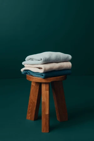 Stack of folded knit sweaters on a wooden stool against a dark teal background. Cozy apparel in a home setting.