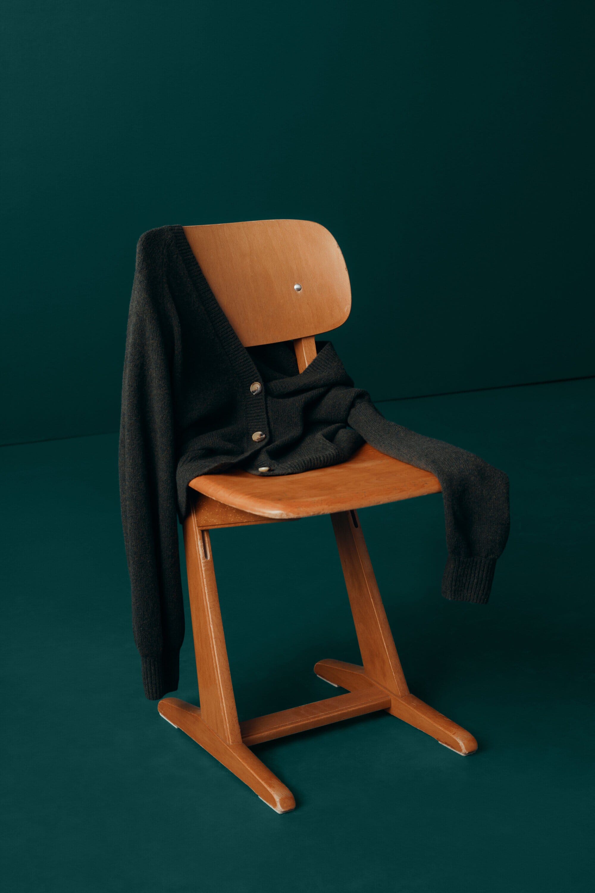A black cardigan is draped over a wooden chair against a dark teal backdrop, creating a minimalist fashion product shot.