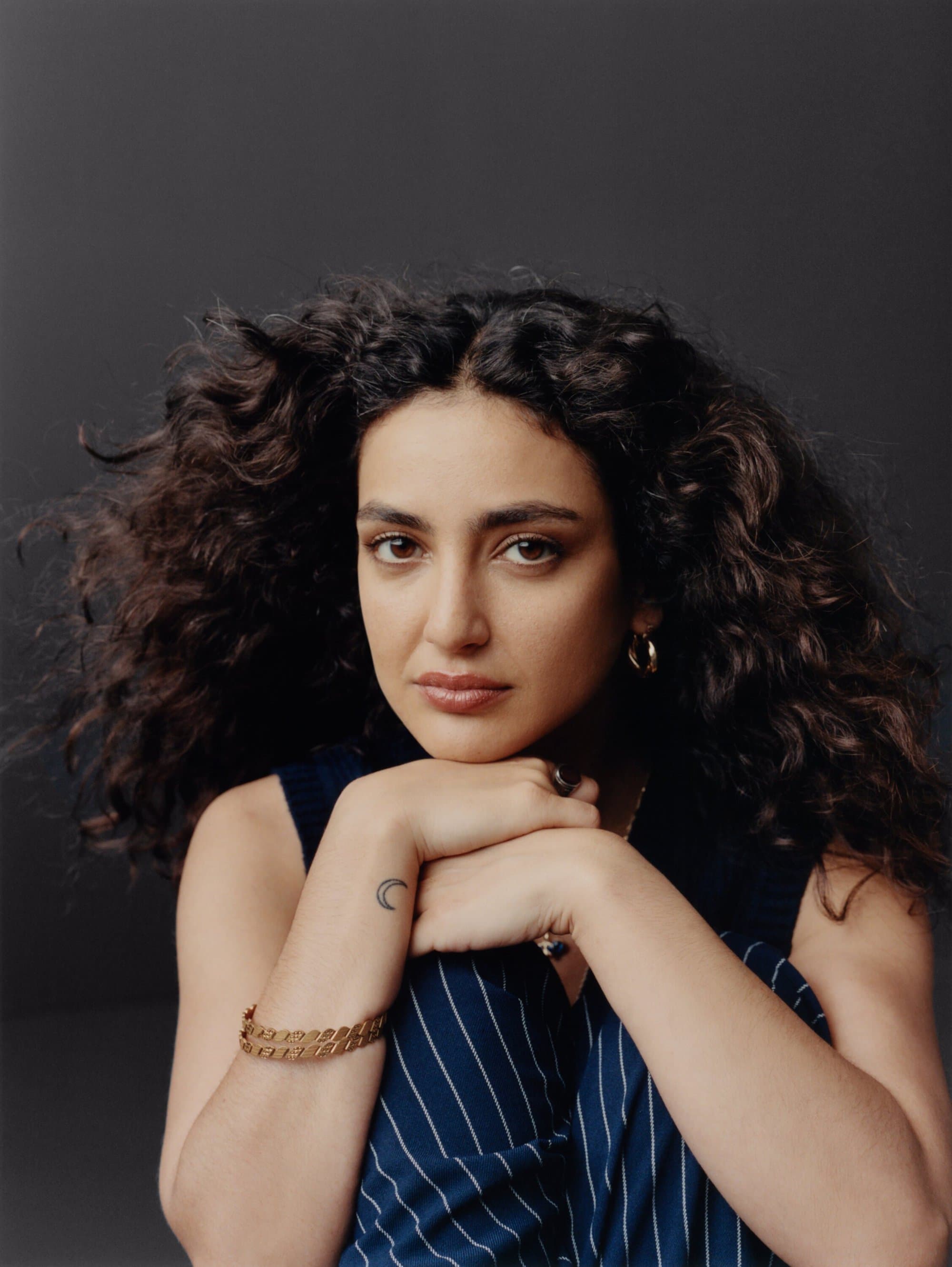 Portrait of a woman with curly hair wearing a blue pinstripe shirt, arms crossed, resting her head on her hands, studio lighting.