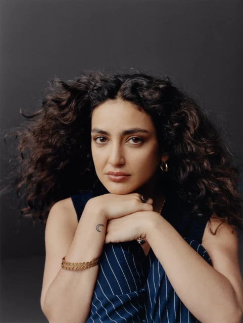 Portrait of a woman with curly hair wearing a blue pinstripe shirt, arms crossed, resting her head on her hands, studio lighting.