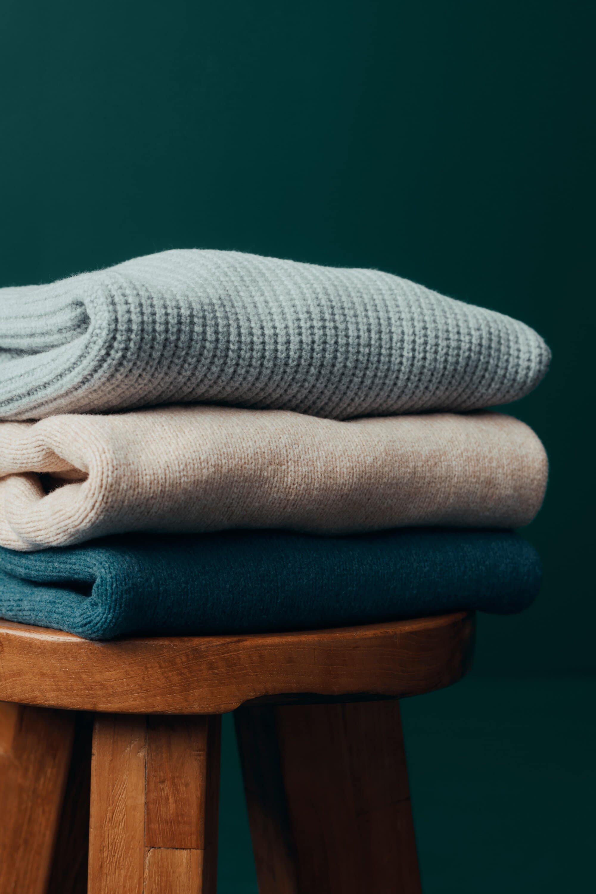 Stack of folded knit sweaters in various colors sits on a wooden stool against a dark green background indoors. Simple, cozy scene.