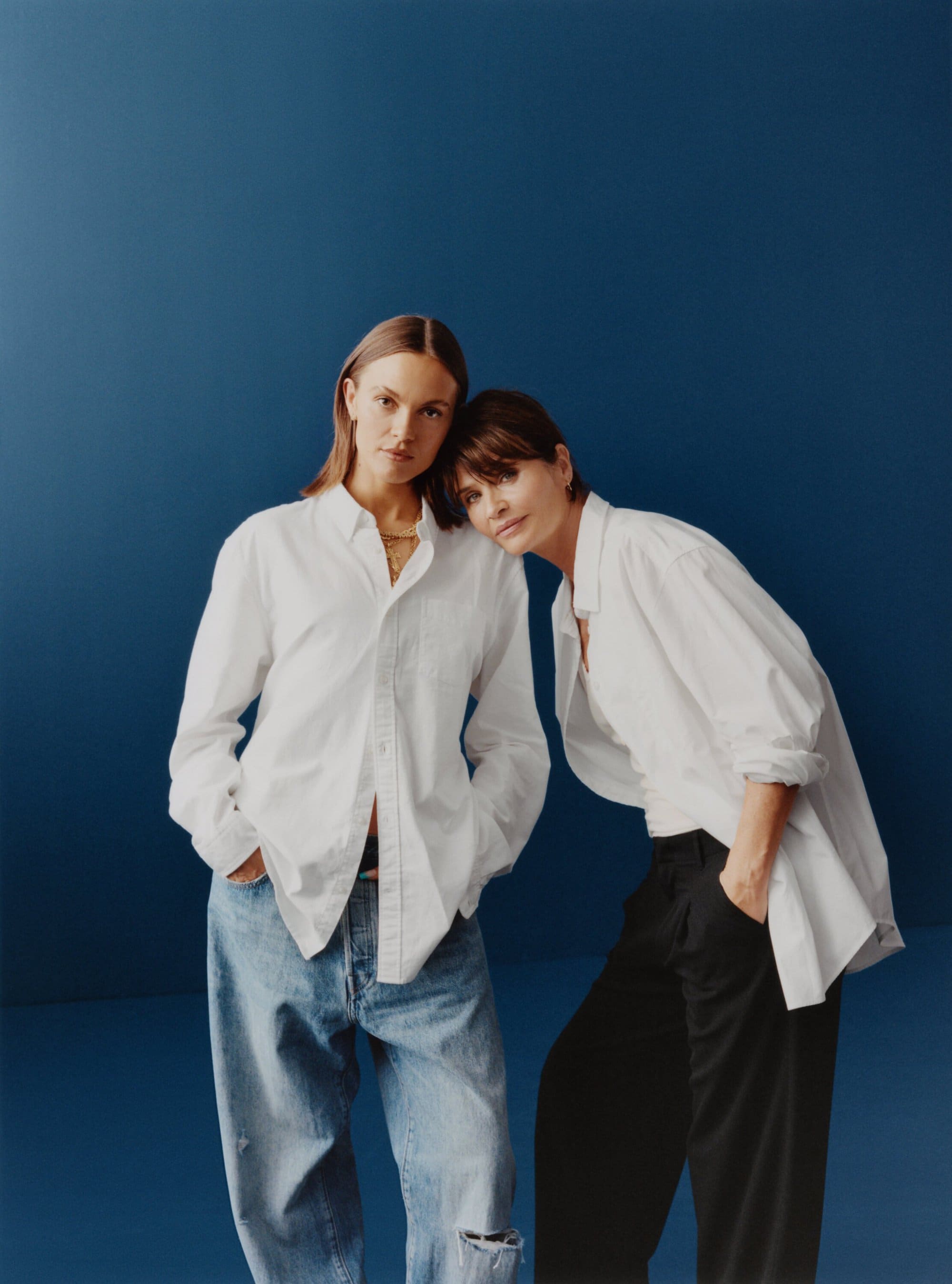 Two women pose in a photo studio against a blue backdrop. One wears jeans, the other dark pants, both wear white shirts and gold necklaces.