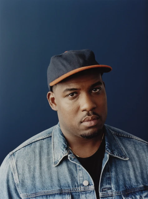 Close-up portrait of an African-American man with a baseball cap and denim jacket, set against a dark blue backdrop in a studio environment.