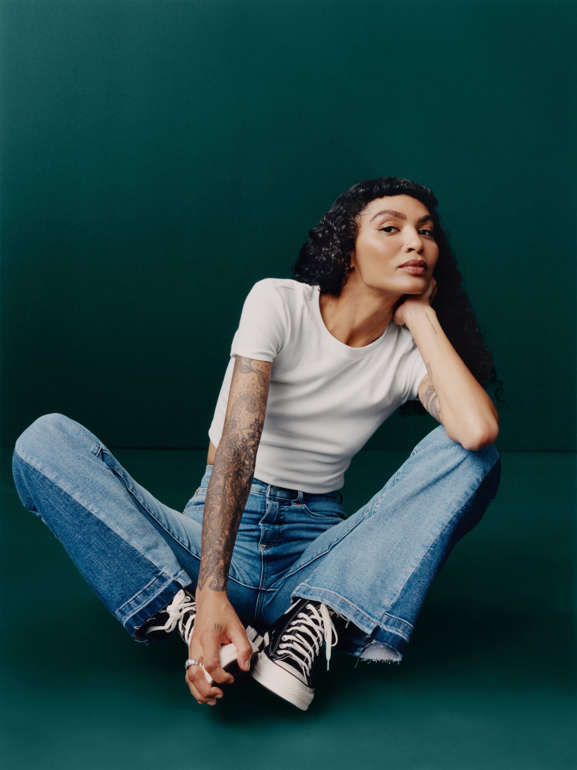 Woman in white top and patchwork jeans poses on a dark green backdrop, showcasing casual fashion with a relaxed, seated posture and dark hair.
