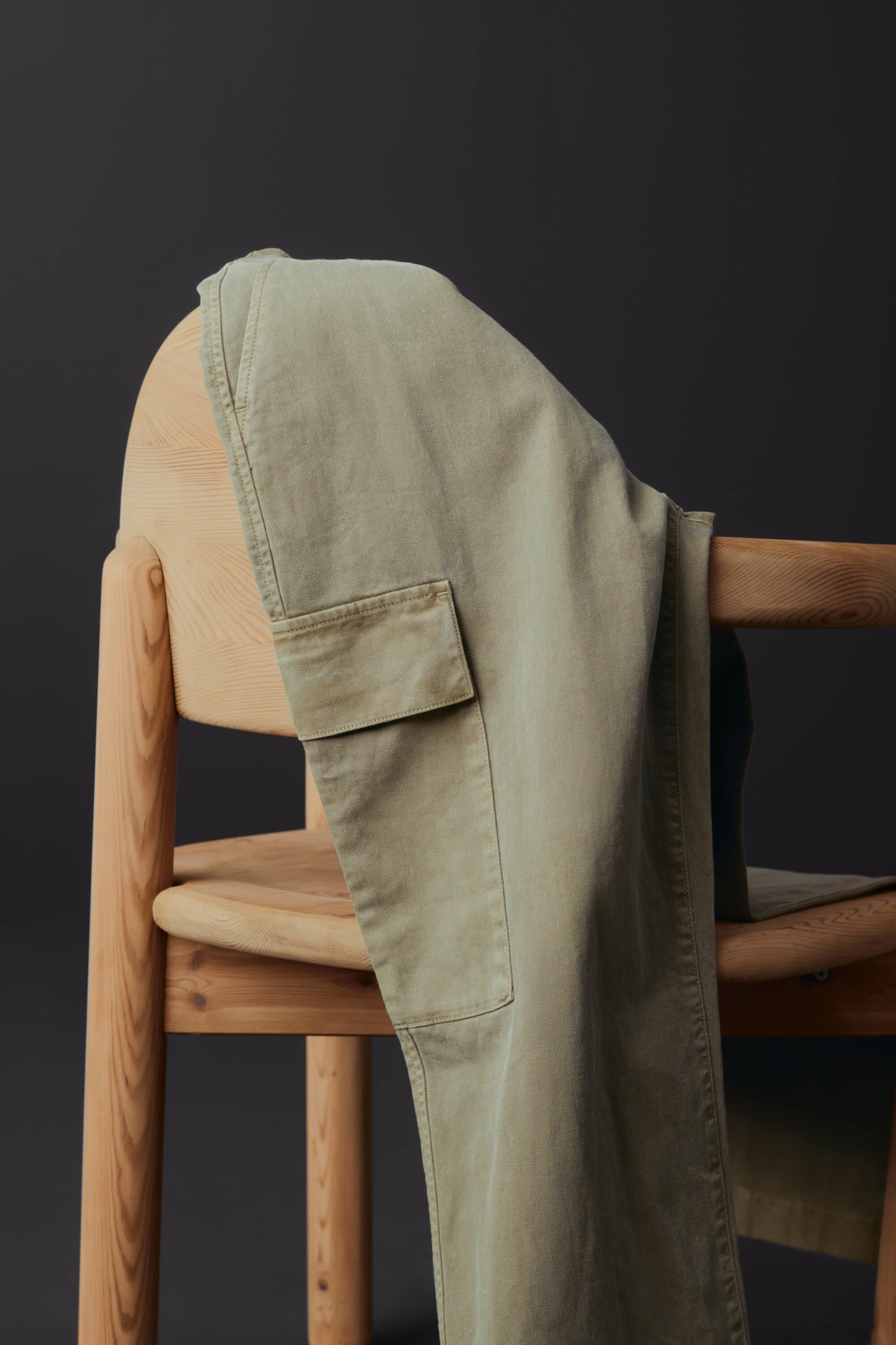 Khaki-colored cargo pants are draped over a light-colored wooden chair against a dark gray background in a studio setting.