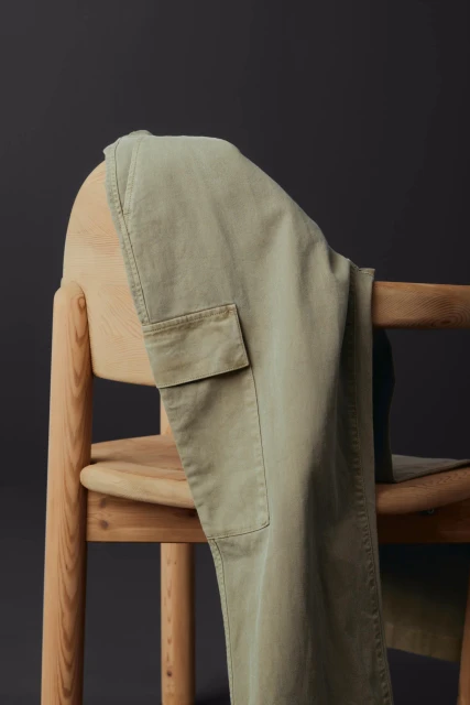 Khaki-colored cargo pants are draped over a light-colored wooden chair against a dark gray background in a studio setting.