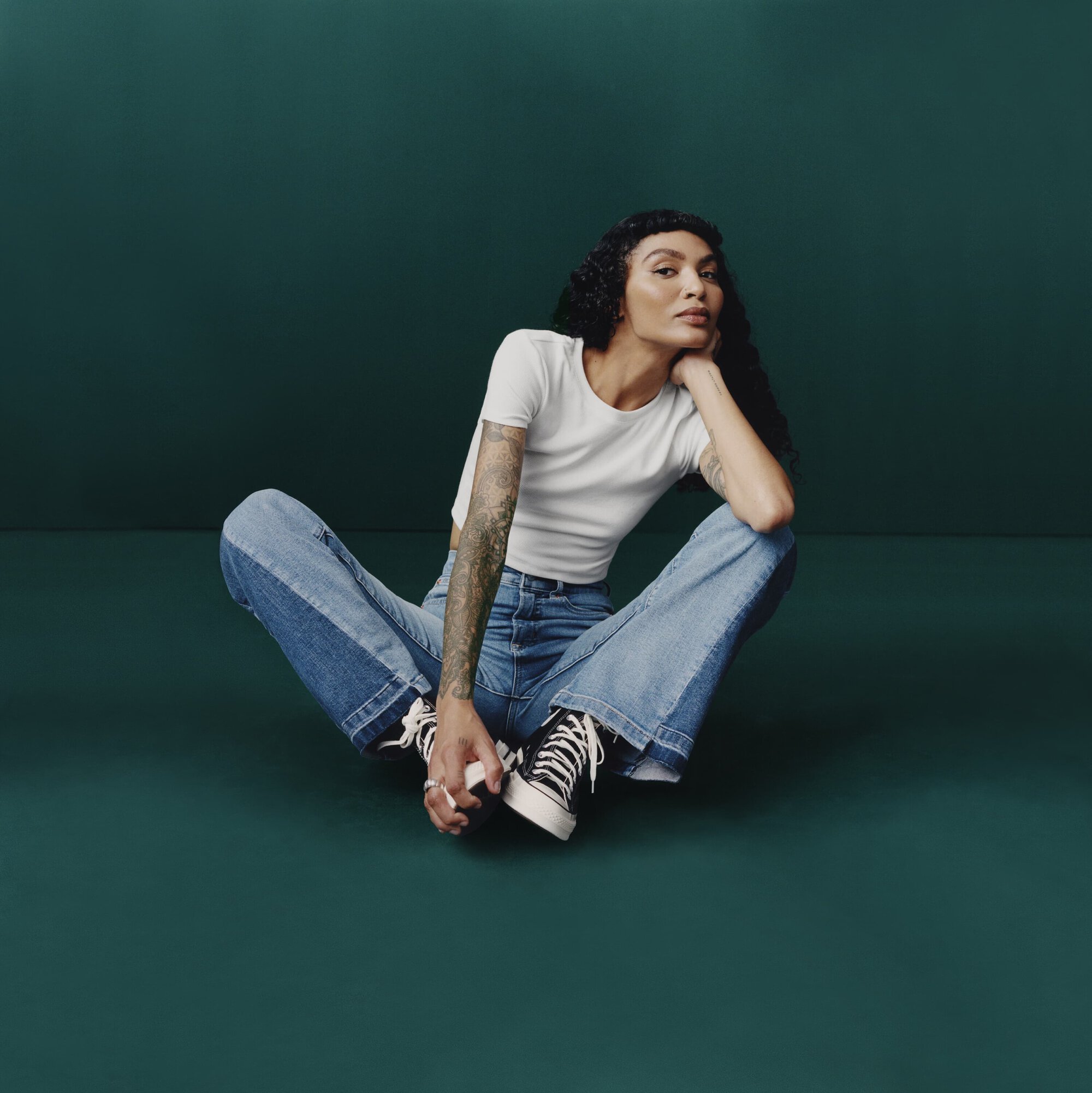 Woman with tattoo sits cross-legged in studio with green background. She wears a white T-shirt, two-tone jeans, and black and white high-tops.