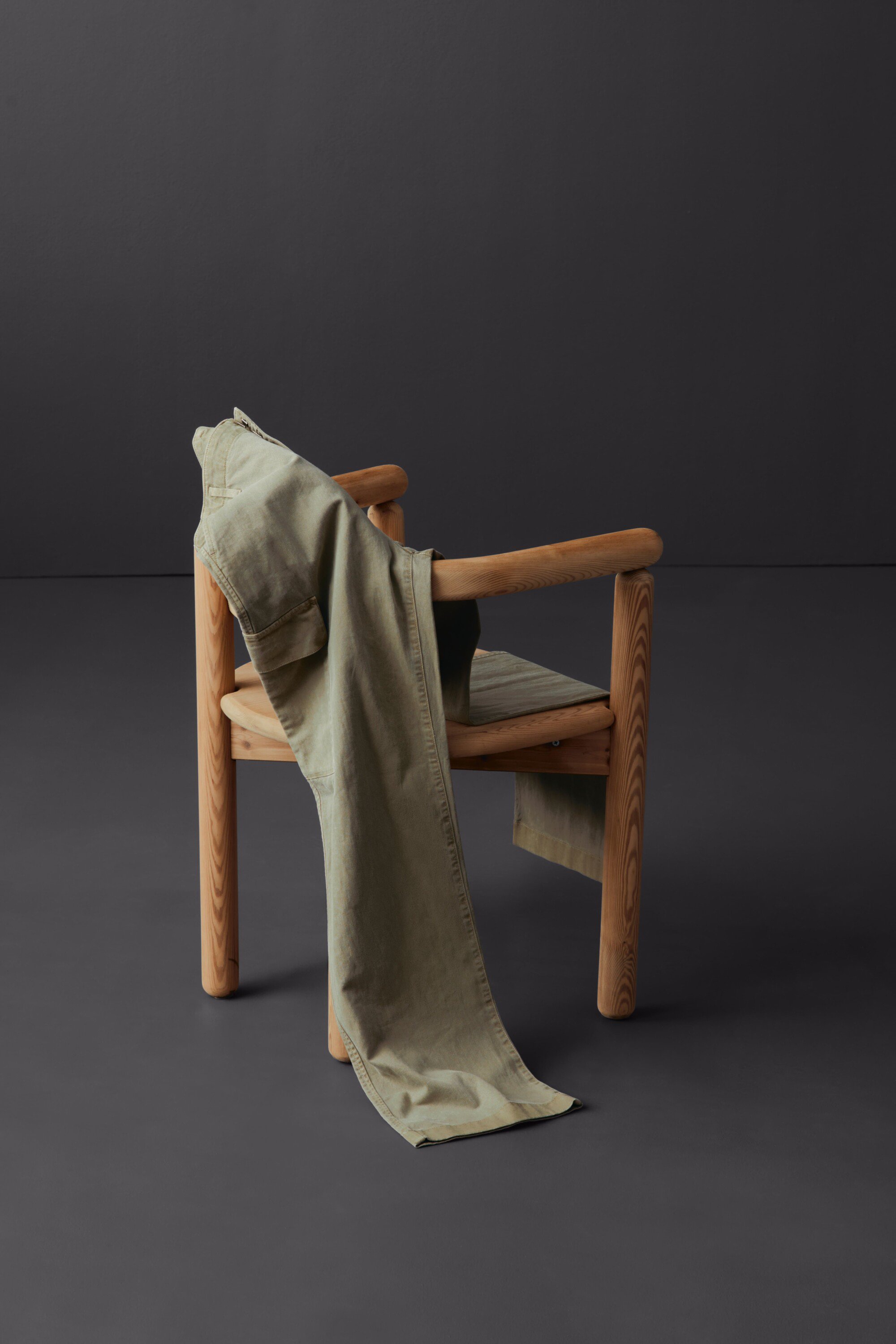 Olive green trousers draped over a light-colored wooden chair against a dark gray background, indoor studio setting, fashion photography.