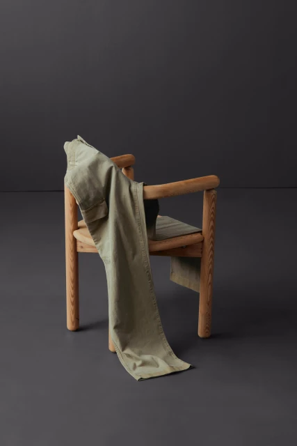 Olive green trousers draped over a light-colored wooden chair against a dark gray background, indoor studio setting, fashion photography.