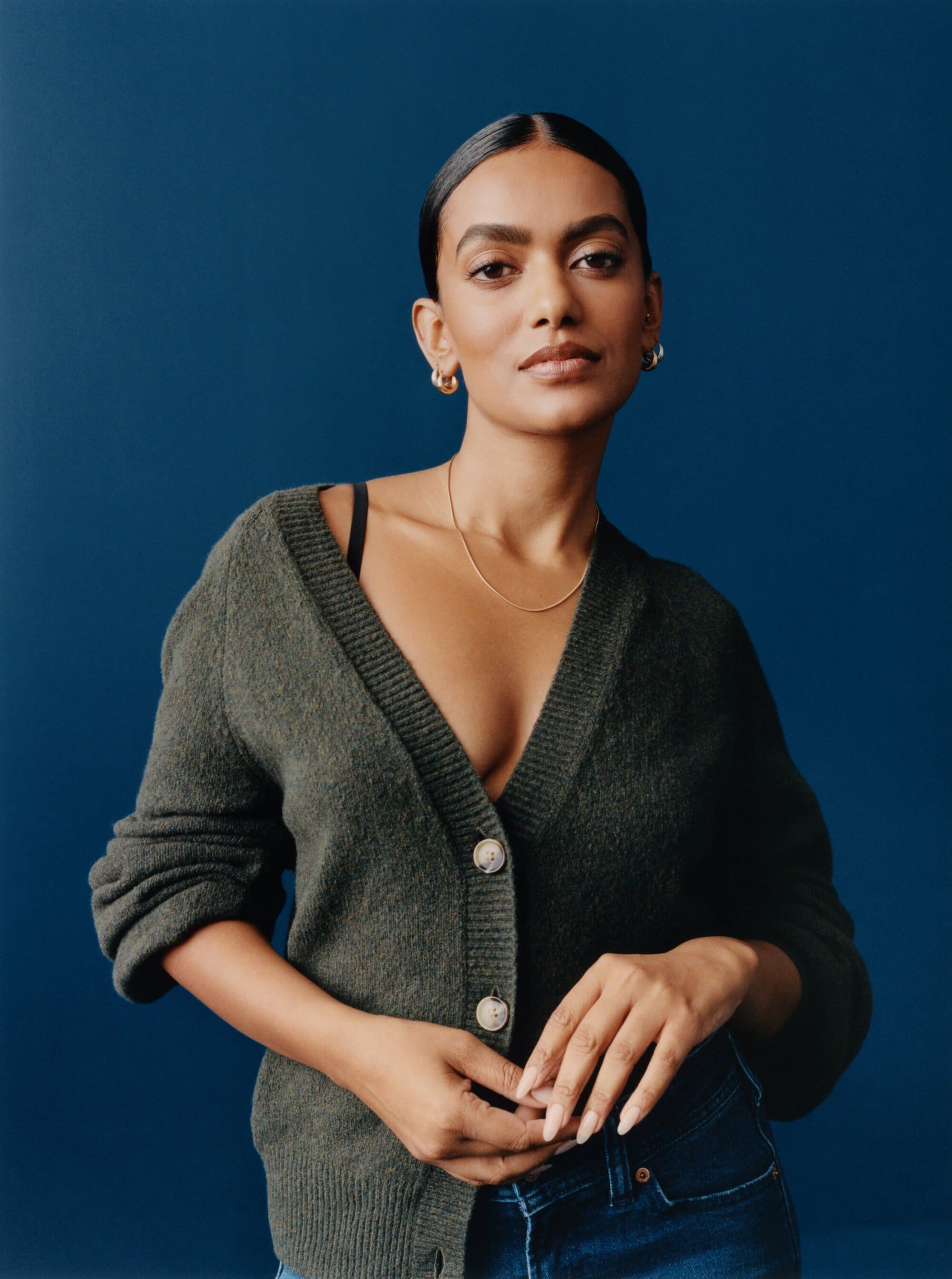 Portrait of a woman wearing a button-down cardigan, gold necklace, and jeans posing in front of a dark blue backdrop.