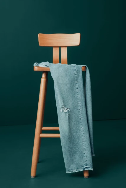 Distressed light wash jeans draped on a light wood stool against a solid dark teal backdrop in a studio setting. Fashion still life.