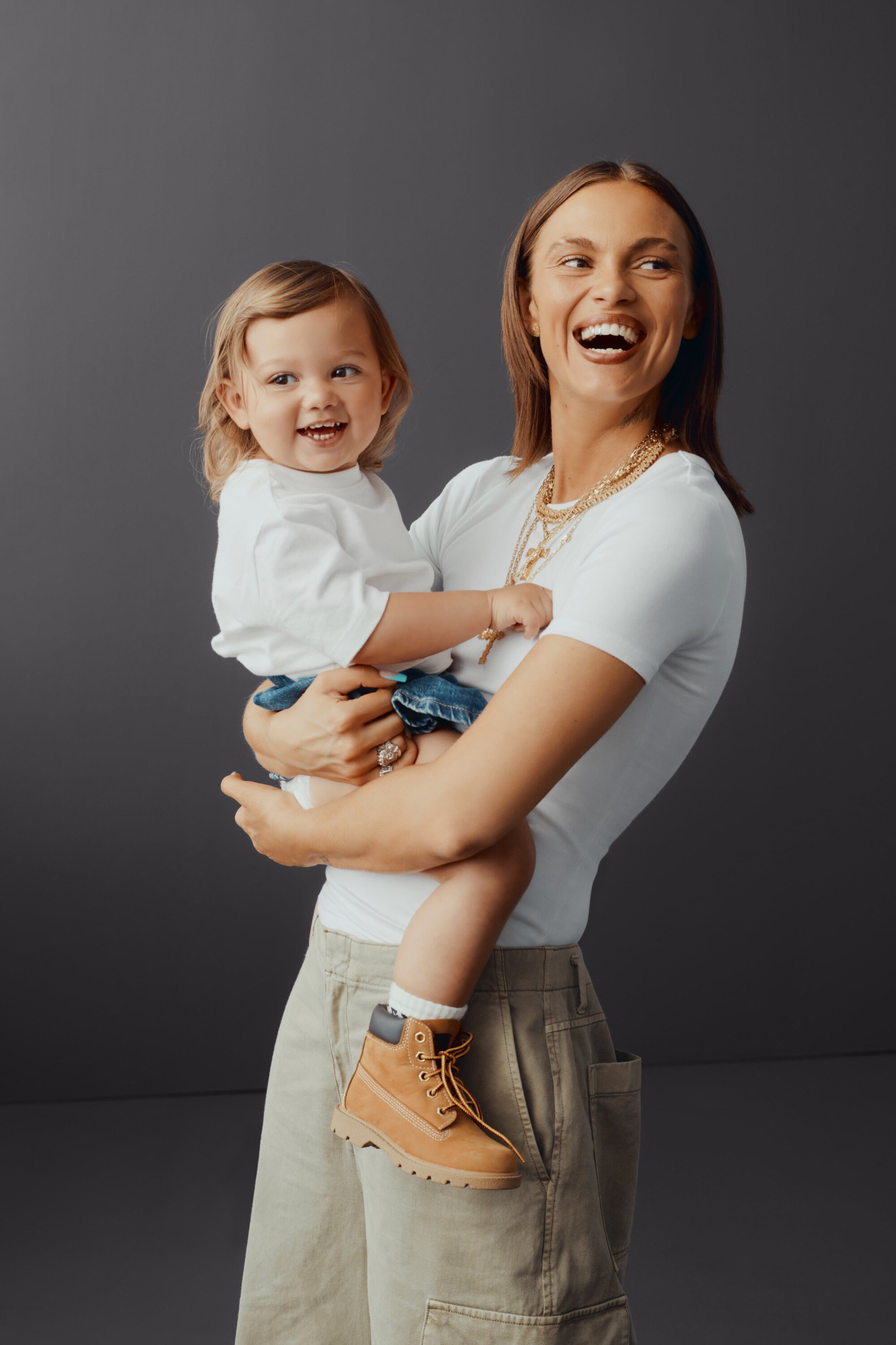 Mother and child share a joyful moment in a studio shot, both smiling and wearing casual attire against a simple grey backdrop.
