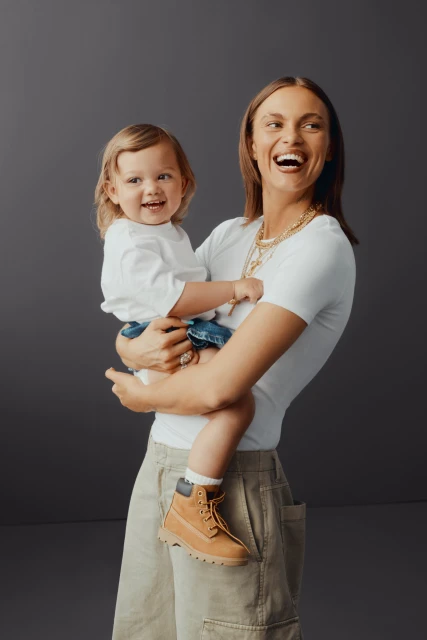 Mother and child share a joyful moment in a studio shot, both smiling and wearing casual attire against a simple grey backdrop.