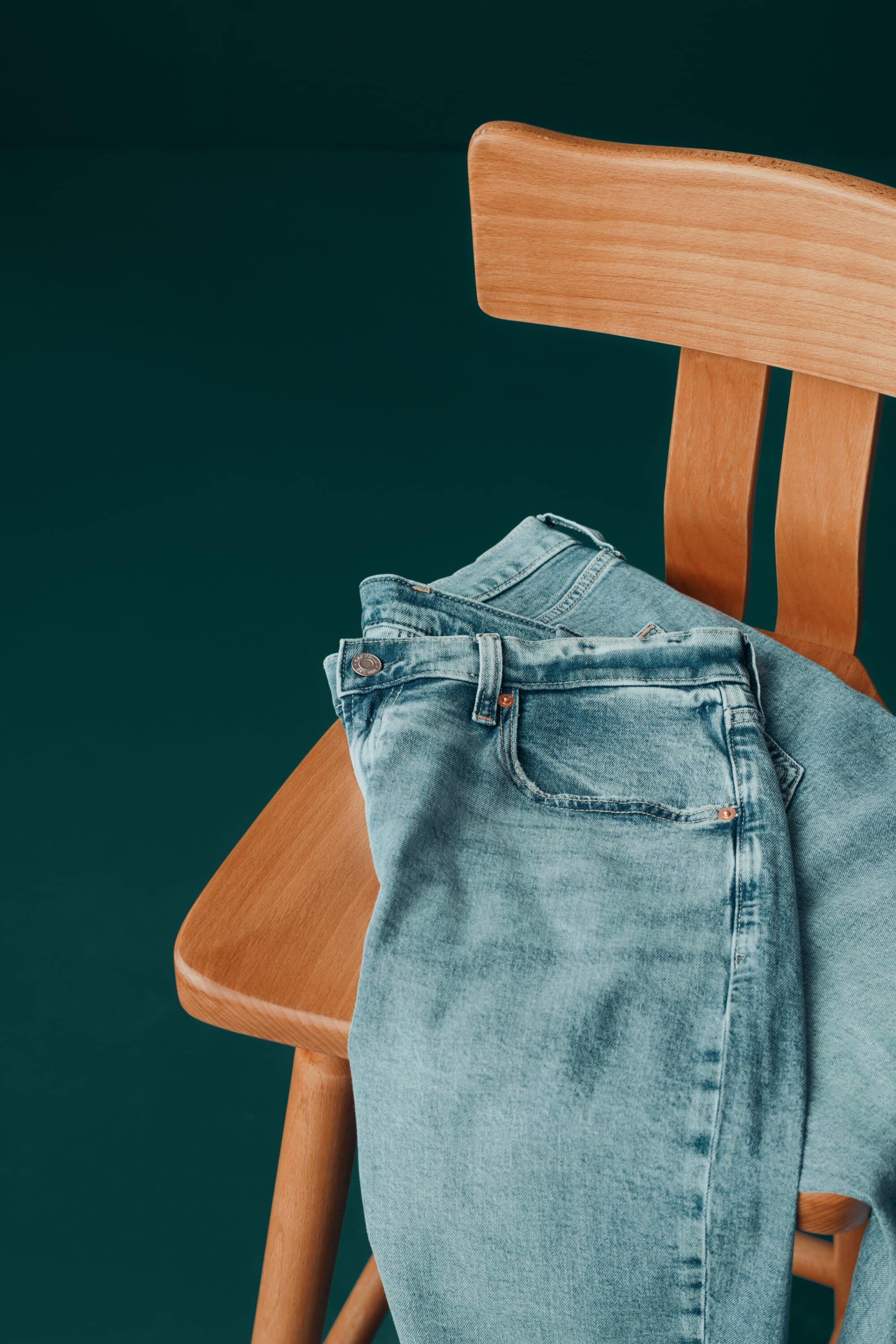 Light blue denim jeans casually draped over a light brown wooden chair against a solid teal background, fashion or clothing advertisement.