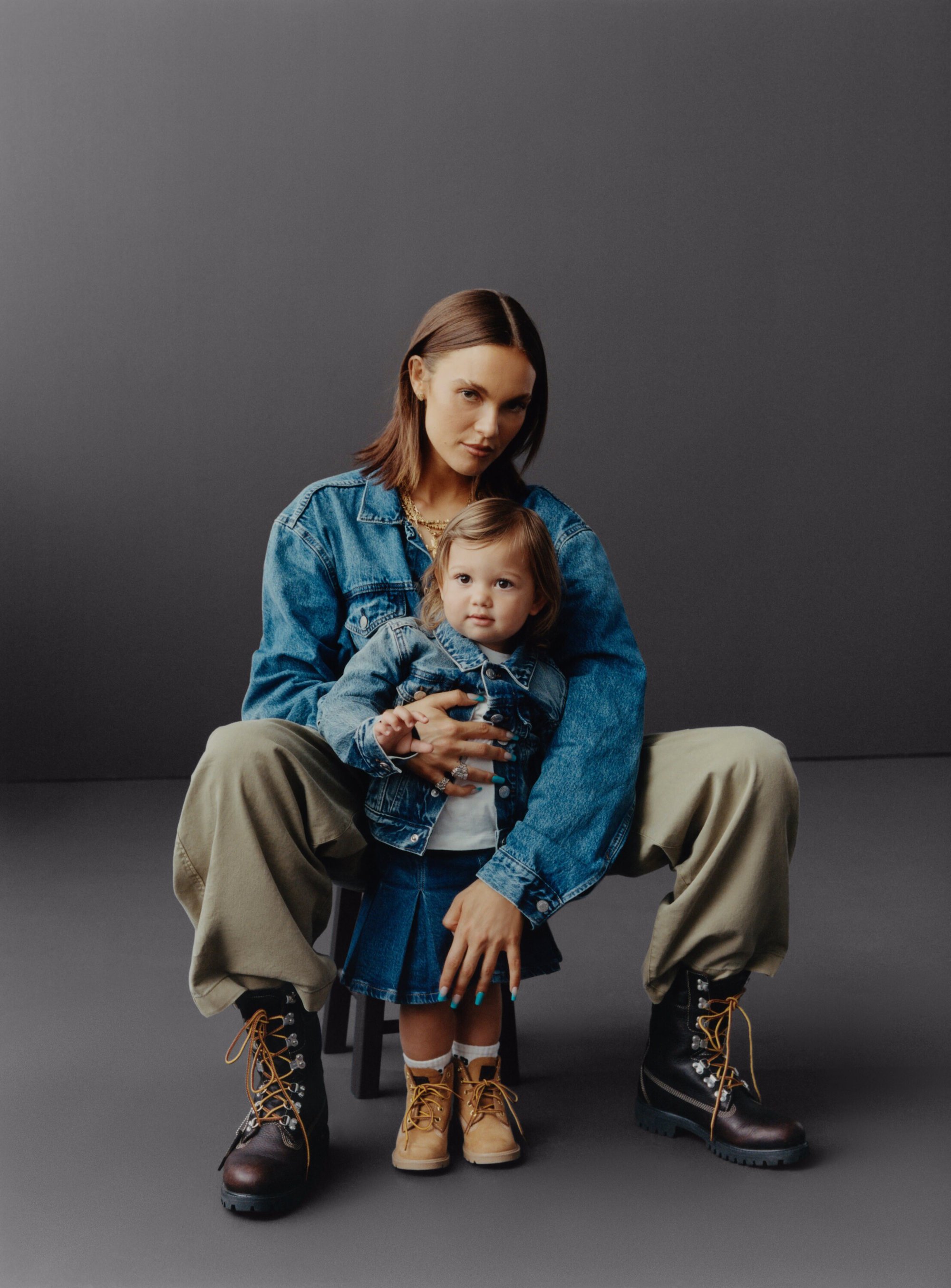 Adult female in a denim jacket seated with a child in matching outfit, both wearing work boots in a photo studio setting.