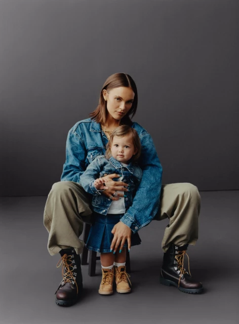 Adult female in a denim jacket seated with a child in matching outfit, both wearing work boots in a photo studio setting.