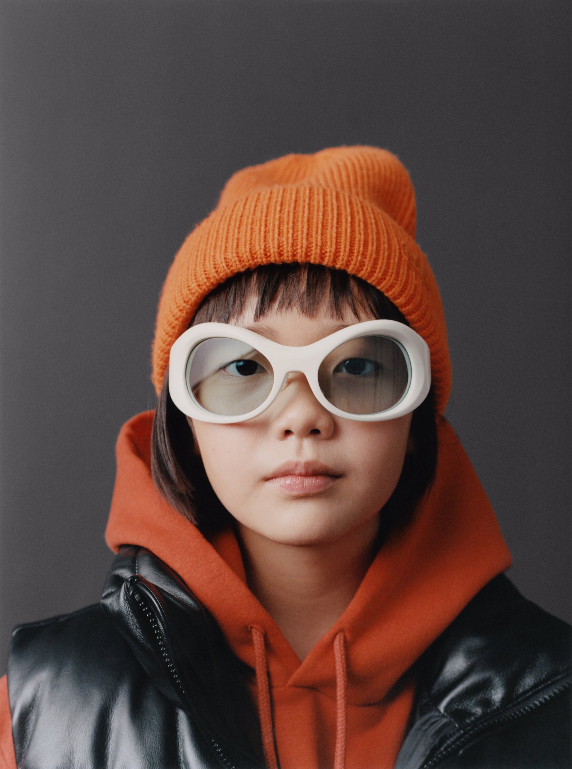 Teen girl portrait in an orange knit cap, black vest, and retro sunglasses against a gray background, fashion portrait, studio shot.