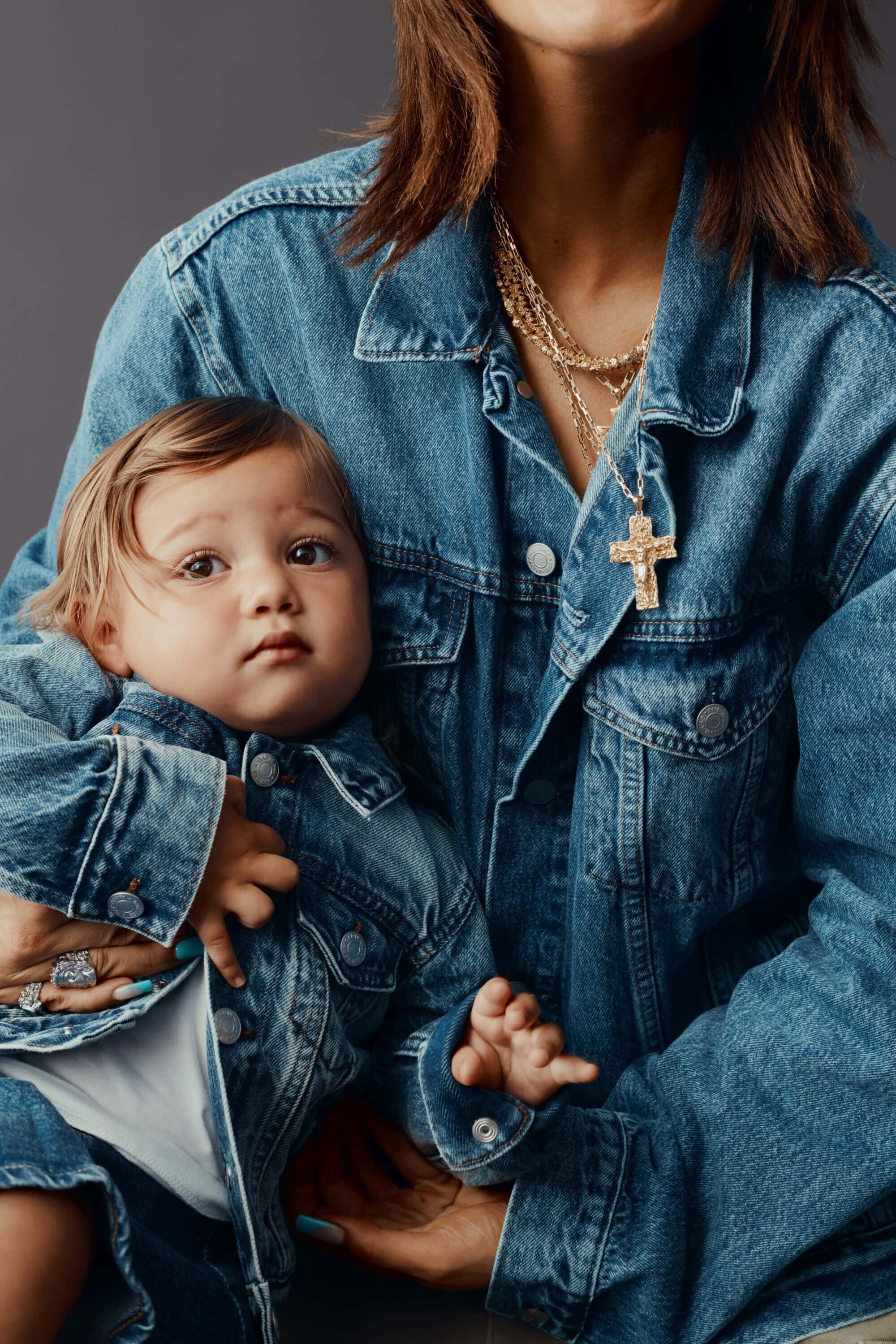 A baby wearing a denim jacket being held by an adult wearing a matching denim jacket and a gold cross necklace indoors.