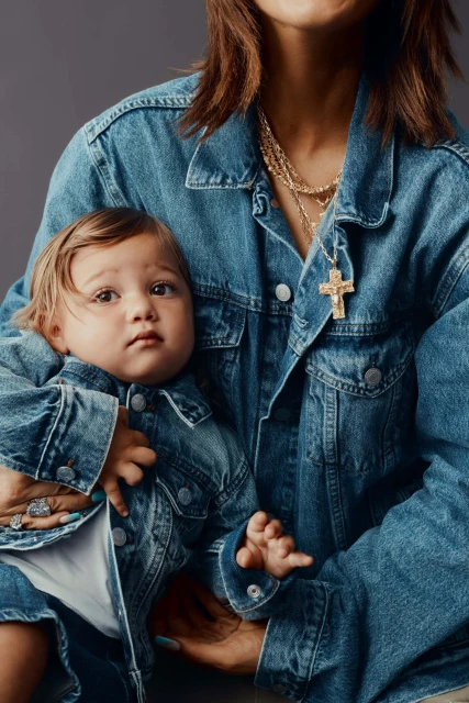 A baby wearing a denim jacket being held by an adult wearing a matching denim jacket and a gold cross necklace indoors.