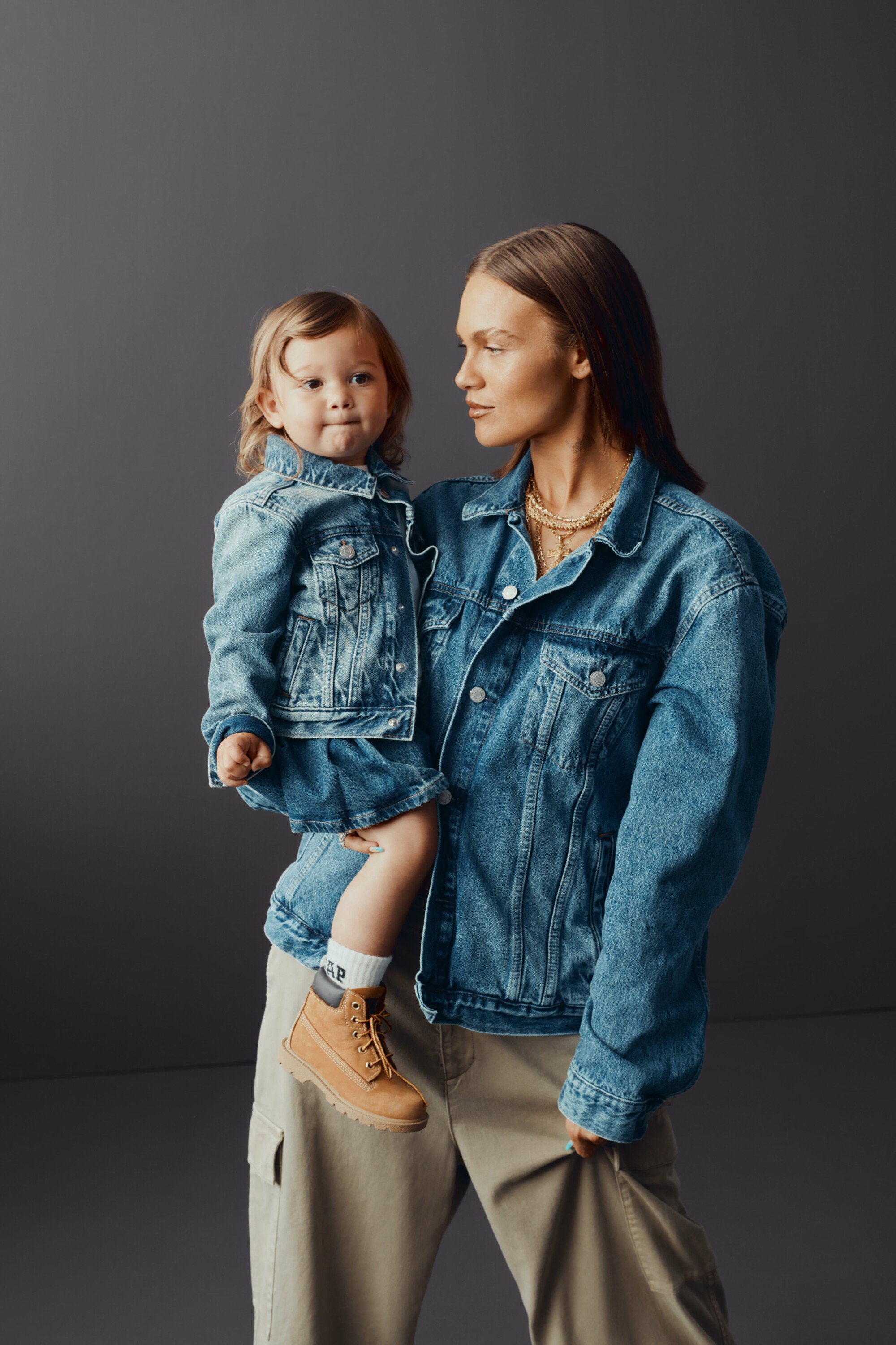 A woman holds a toddler, both wearing denim jackets and tan boots, standing indoors against a grey background in a portrait-style shot.