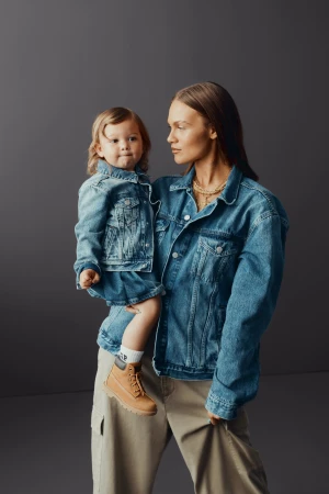 A woman holds a toddler, both wearing denim jackets and tan boots, standing indoors against a grey background in a portrait-style shot.