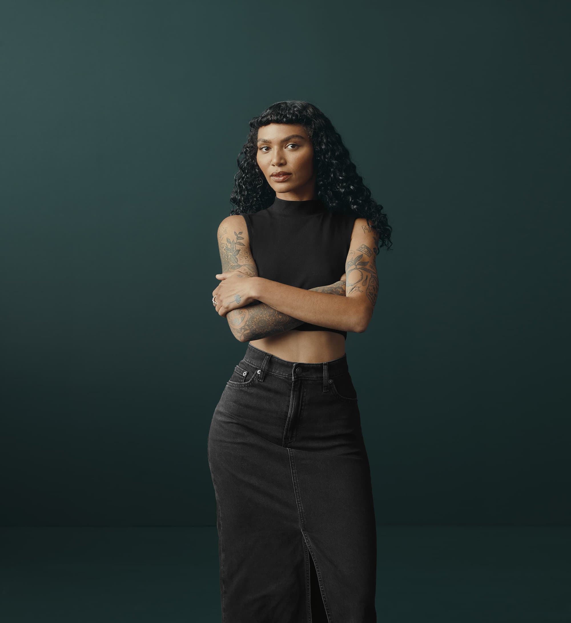 Portrait of an adult woman with curly dark hair wearing a black top and long black skirt, arms crossed in front of a dark green background.