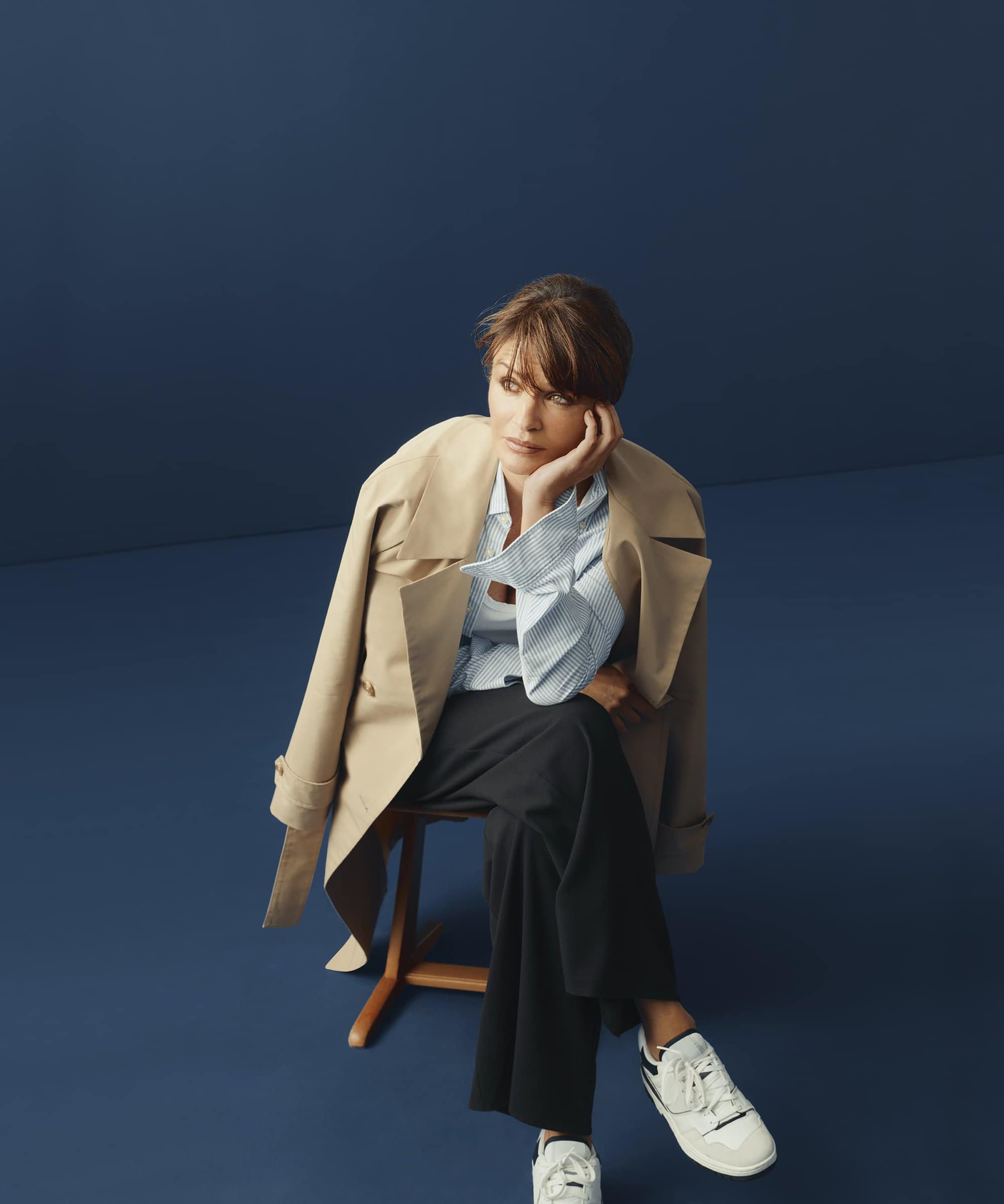 A young woman with short hair in a beige trench coat and black pants sits on a chair against a dark blue backdrop in a fashion portrait.