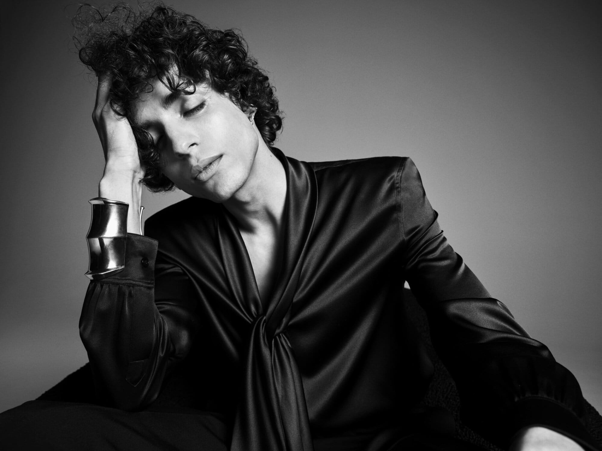Black-and-white portrait of a young adult male model with curly hair, eyes closed, wearing a silky shirt and a large cuff bracelet.