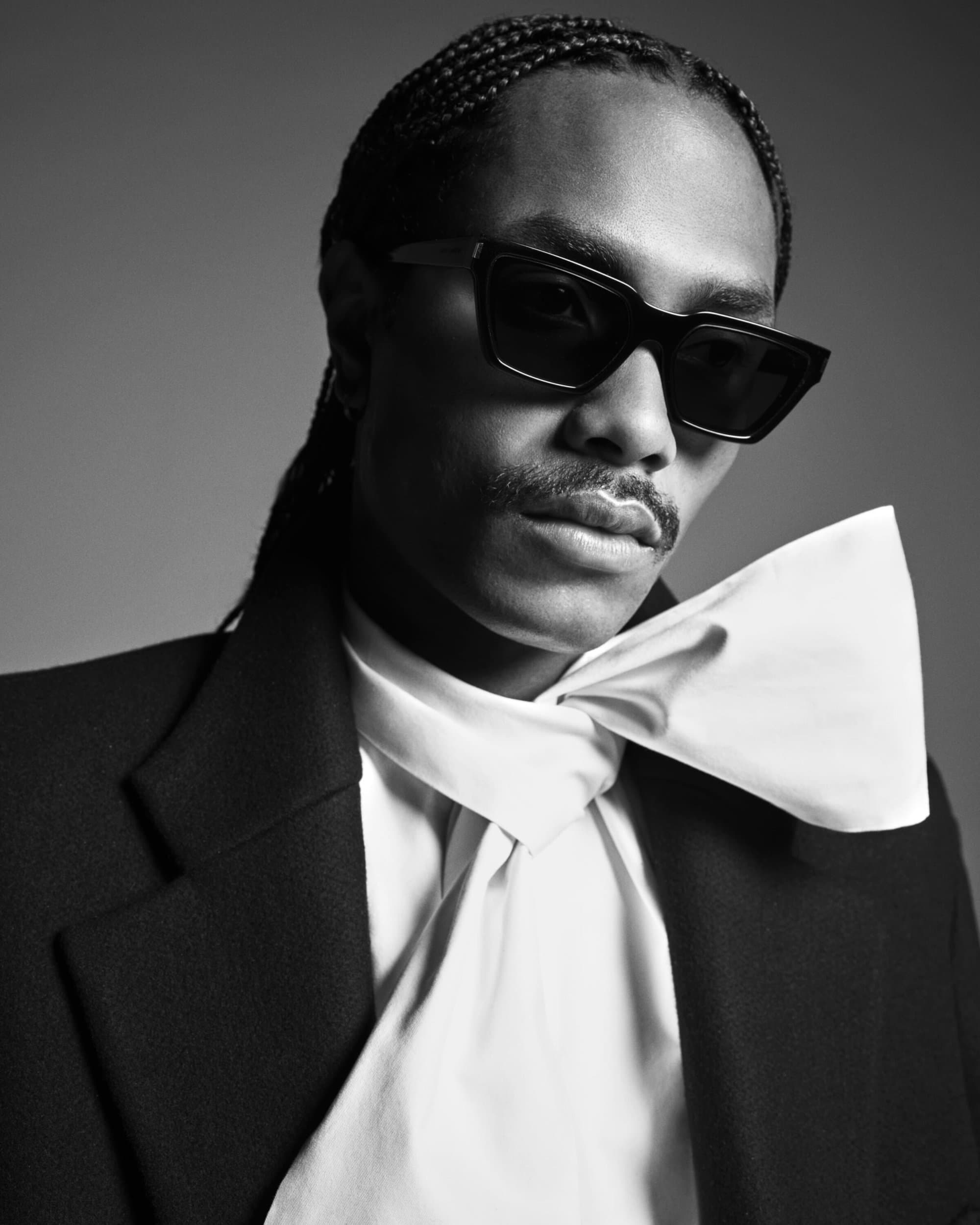 Monochrome portrait of stylish adult male with braided hair, sunglasses, blazer, and large bow tie in a studio setting.