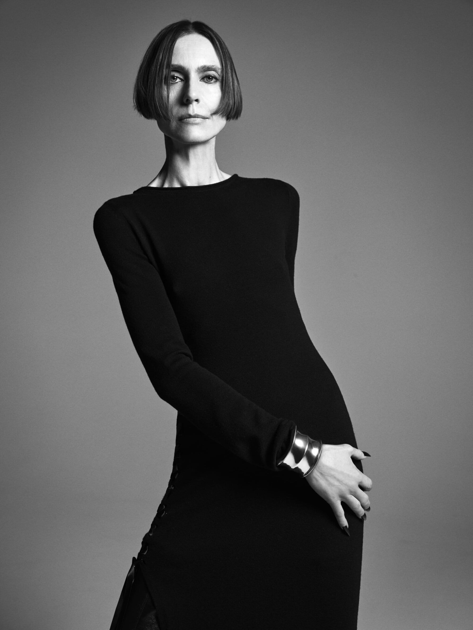 Woman posing in black dress with silver cuff in photo studio. Portrait showcases sleek, modern fashion against neutral backdrop.