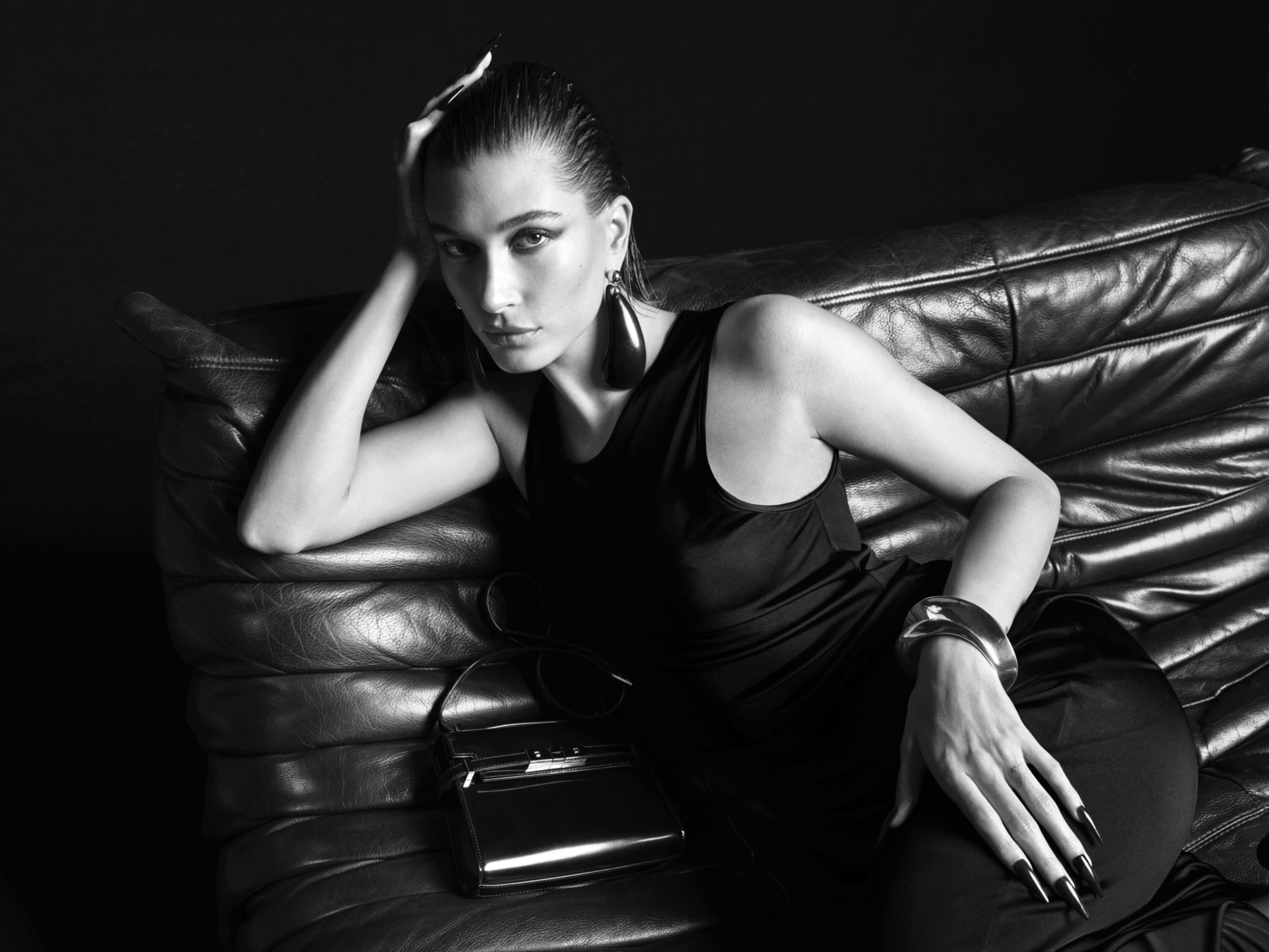 Monochrome portrait of a woman with wet hair and statement jewelry, reclining on a leather sofa with a handbag.