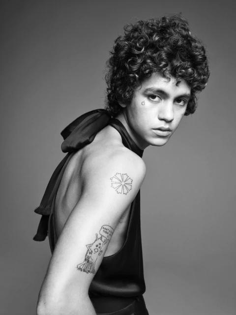 Black and white portrait of a young adult with curly hair and tattoos, wearing a draped sleeveless top against a grey background.