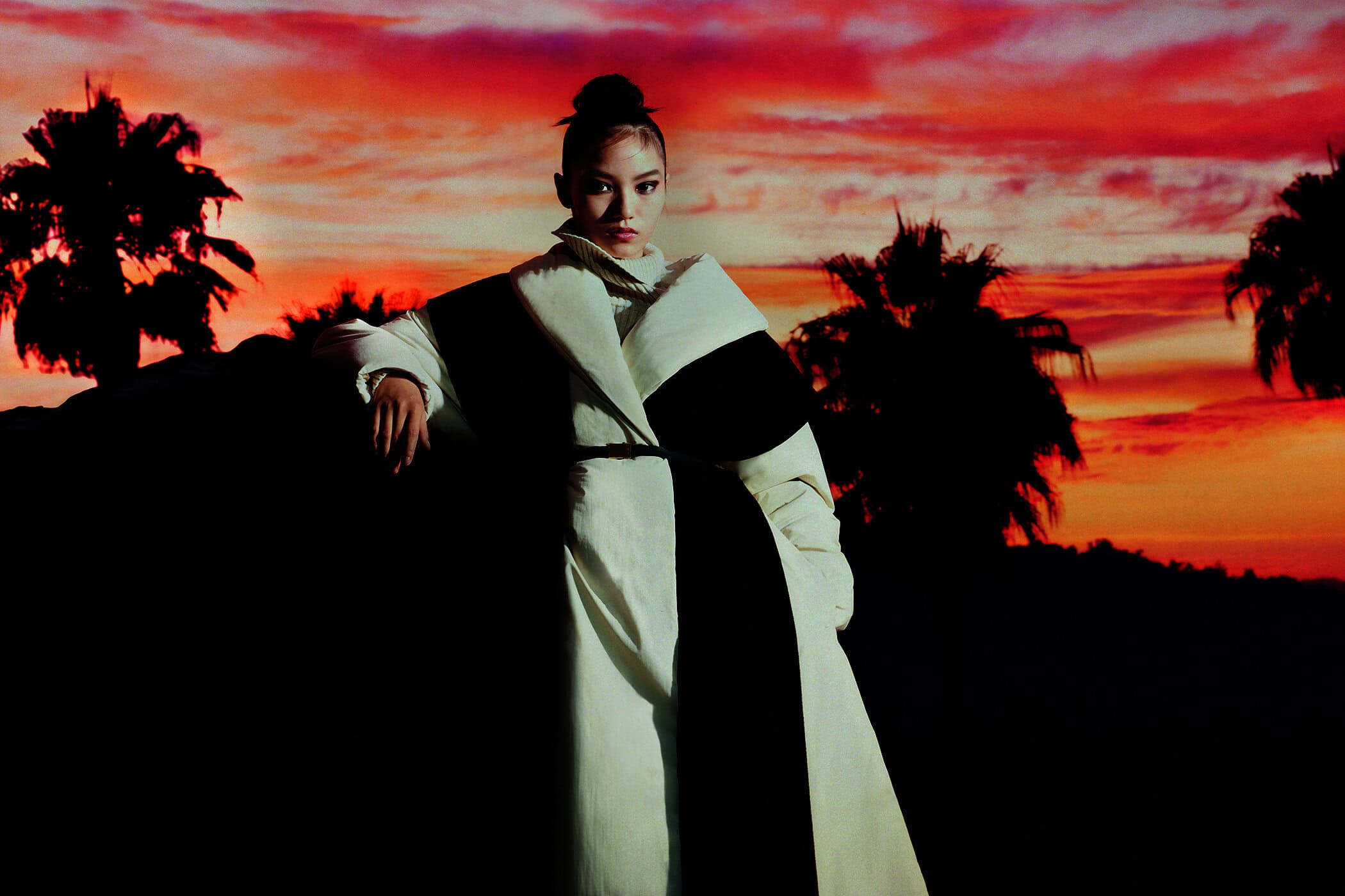 A woman in a white coat poses against a backdrop of palm trees silhouetted by a vibrant orange and red sunset sky.