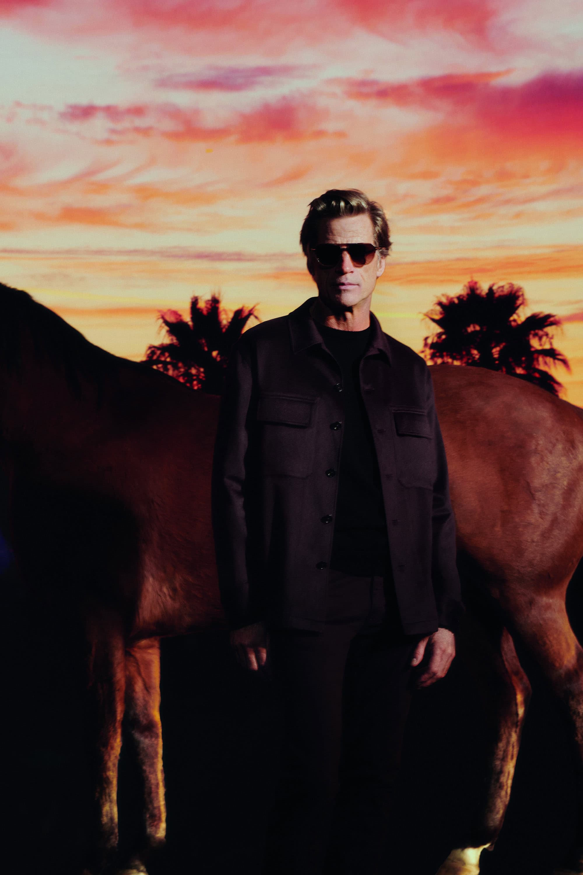 A man in sunglasses and a dark jacket poses next to a brown horse under a colorful sky with palm trees at sunset.