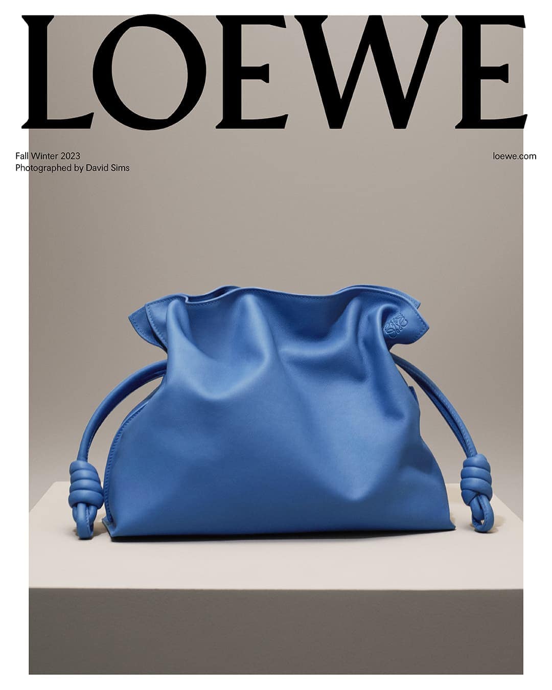 Blue Loewe bag sits on a gray platform against a gray backdrop with Loewe branding and text overlay for a Fall/Winter 2023 fashion advertisement.