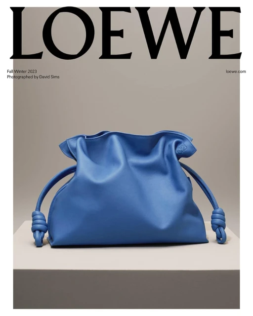 Blue Loewe bag sits on a gray platform against a gray backdrop with Loewe branding and text overlay for a Fall/Winter 2023 fashion advertisement.