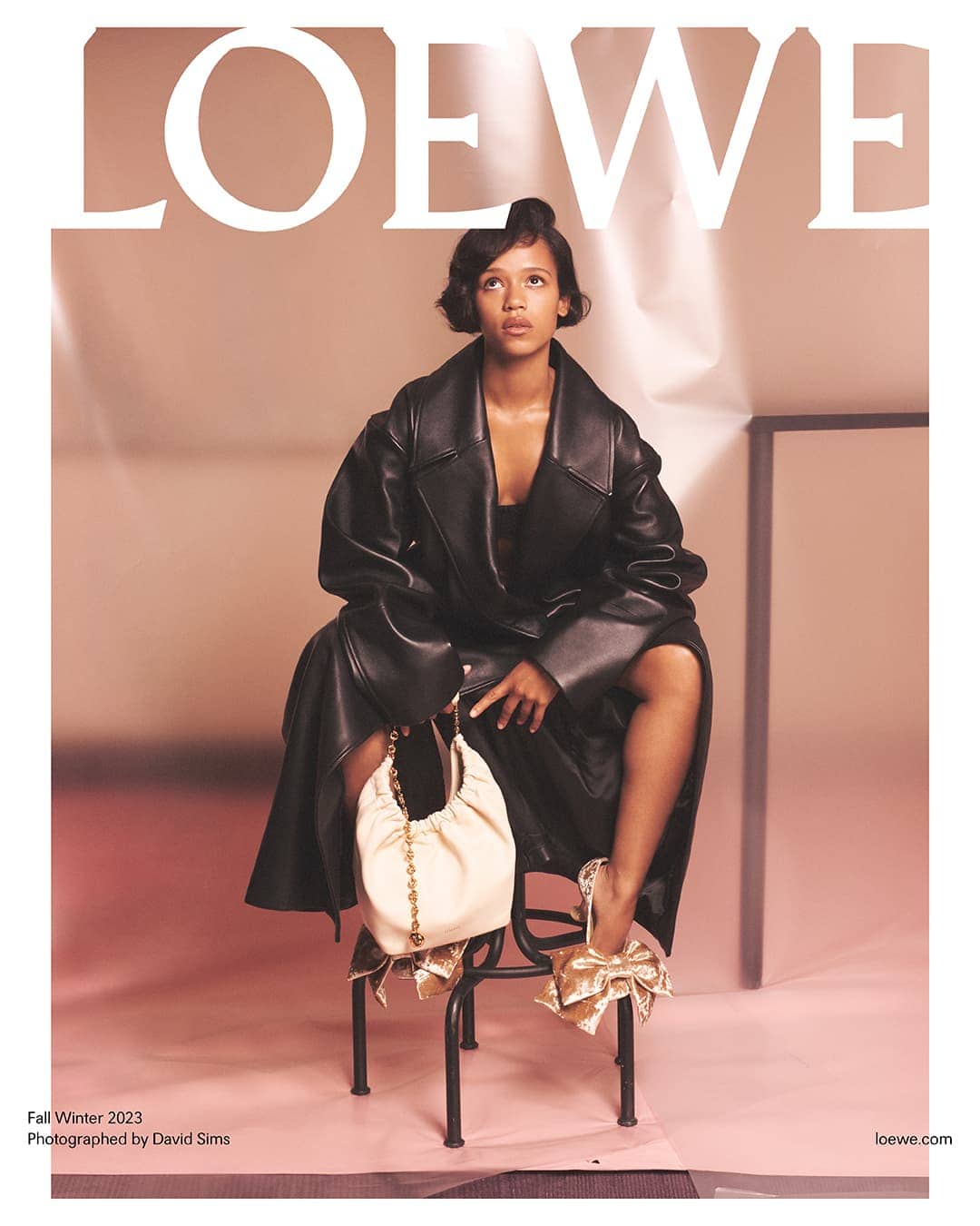Fashion editorial featuring a woman in a black leather coat, sitting on a metal chair, holding a cream-colored bag in a studio setting.