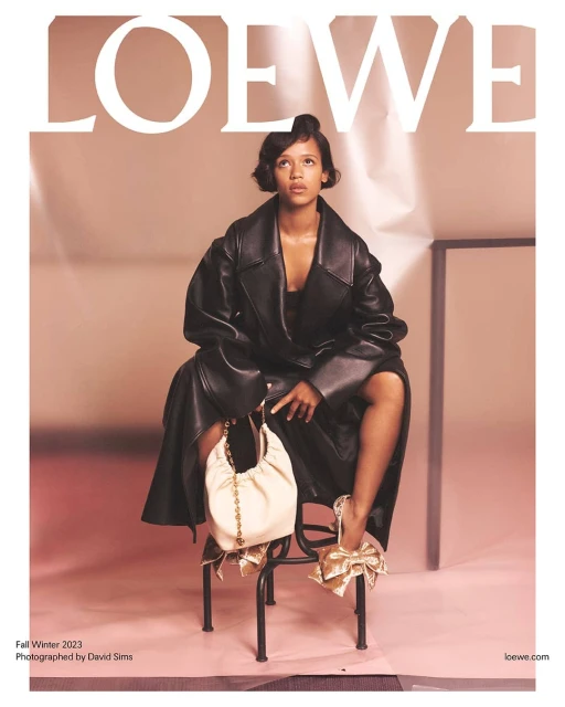 Fashion editorial featuring a woman in a black leather coat, sitting on a metal chair, holding a cream-colored bag in a studio setting.